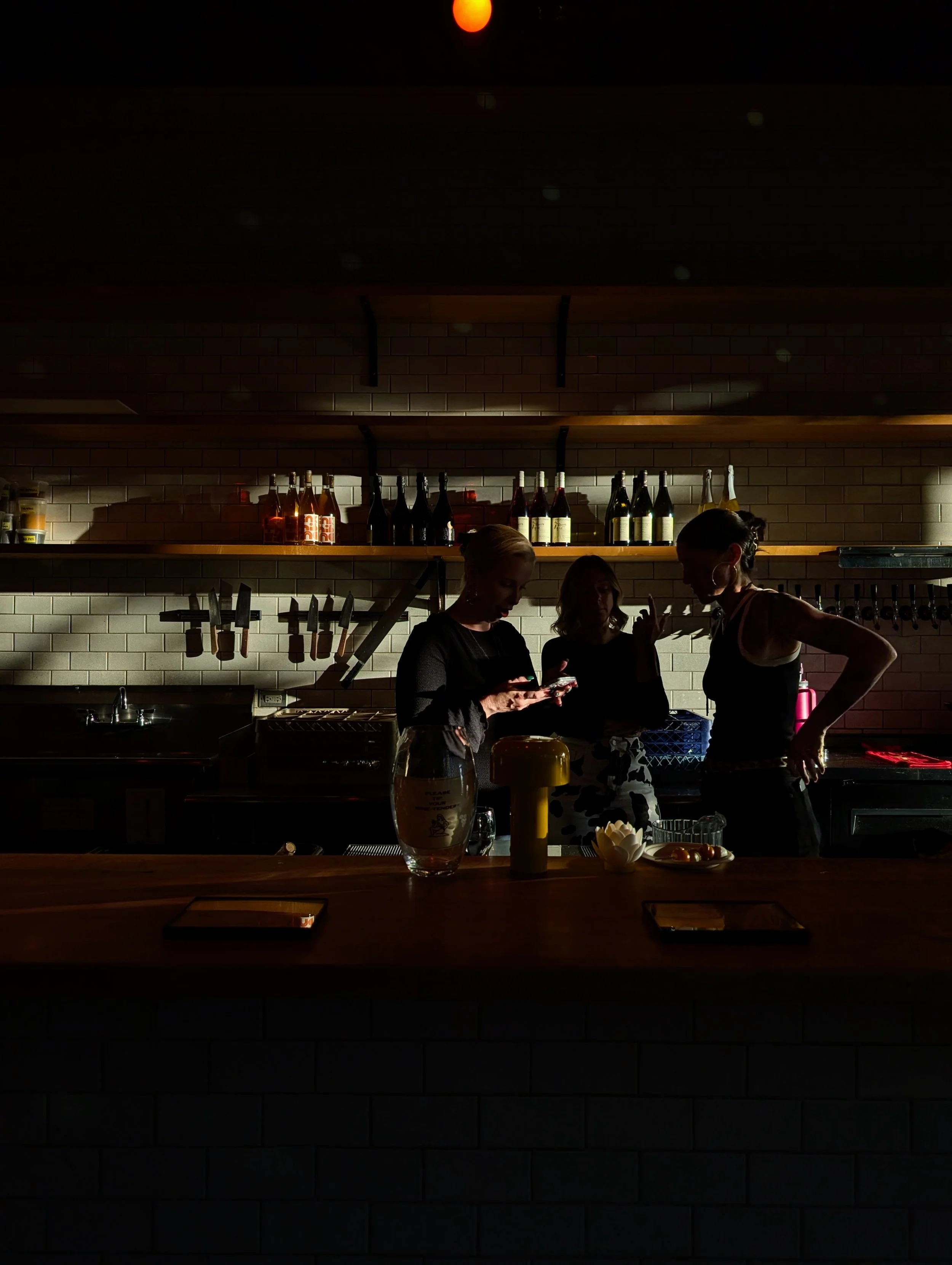 Three women gathered behind a bar in a dimly lit setting, with bottles of alcohol on shelves and knives on display in the background.