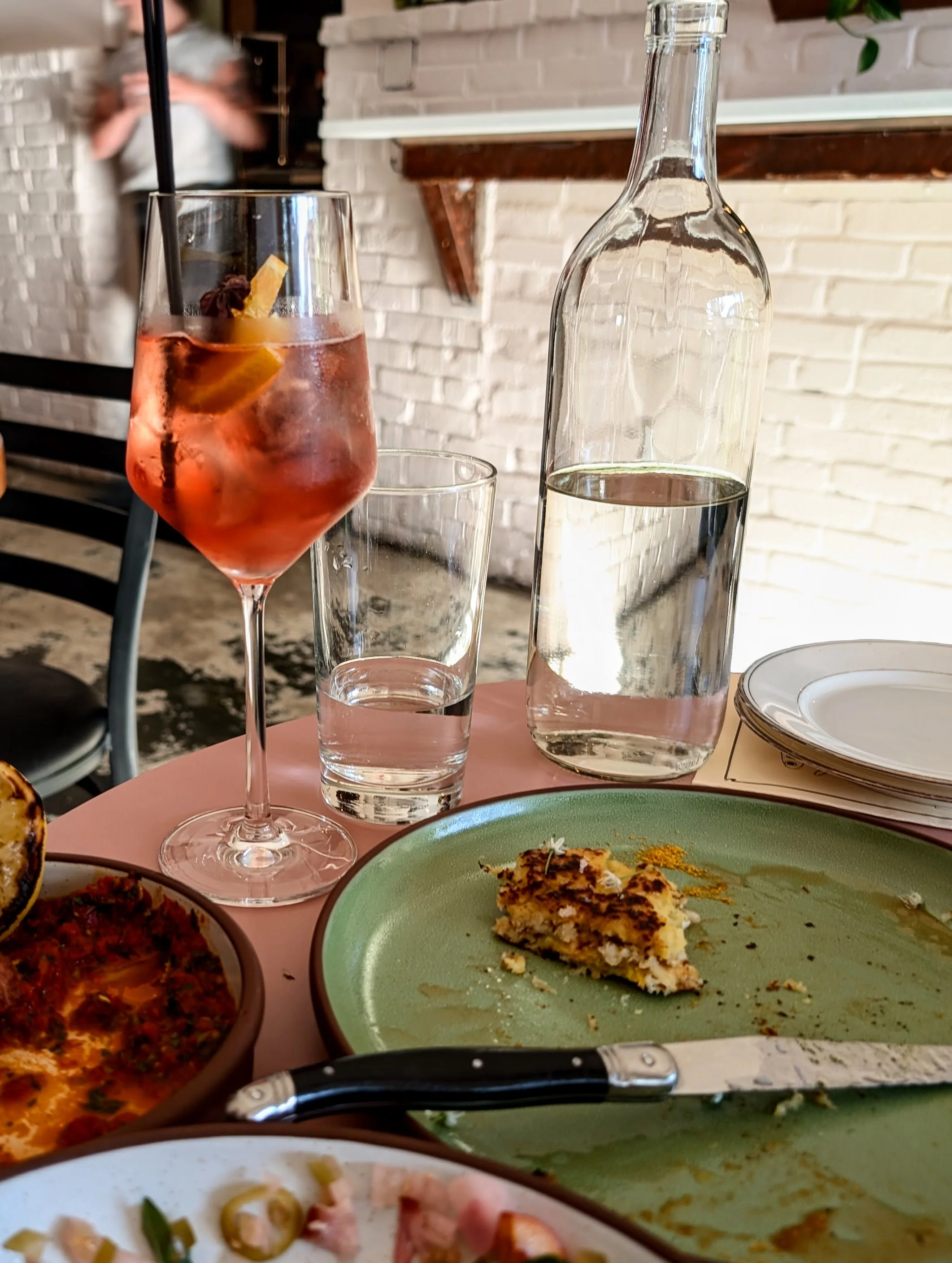 Partially eaten meal on green and white plates with a knife, a half-eaten dish, a glass of red drink with a lemon slice and straw, a glass of water, a bottle of water, and empty plates on a pink table in a restaurant.