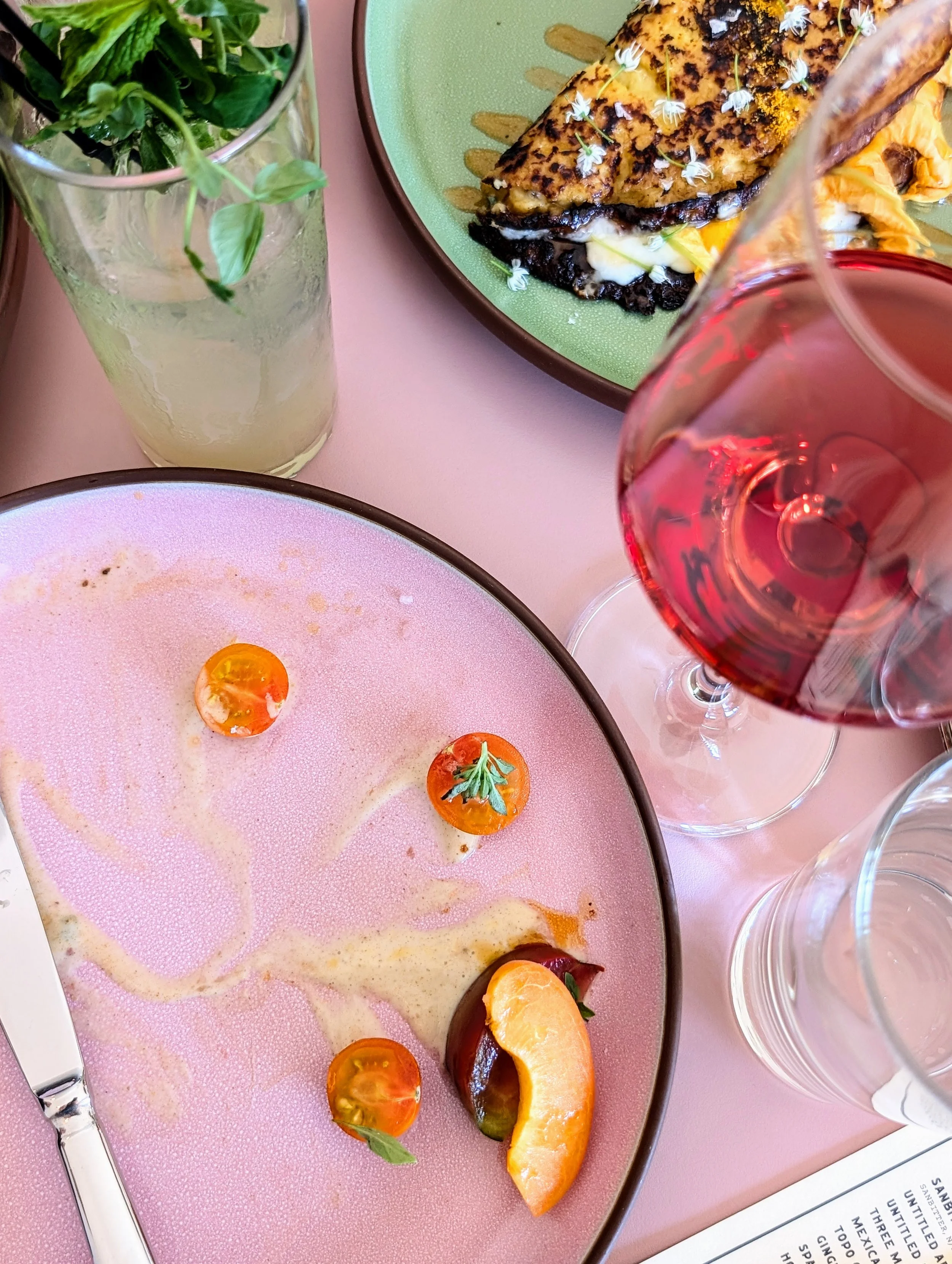 A table setting with a pink plate of cheese, cherry tomatoes, a cantaloupe slice, and a knife, a glass of red wine, a glass of water with mint, and a green plate with grilled fish and vegetables.
