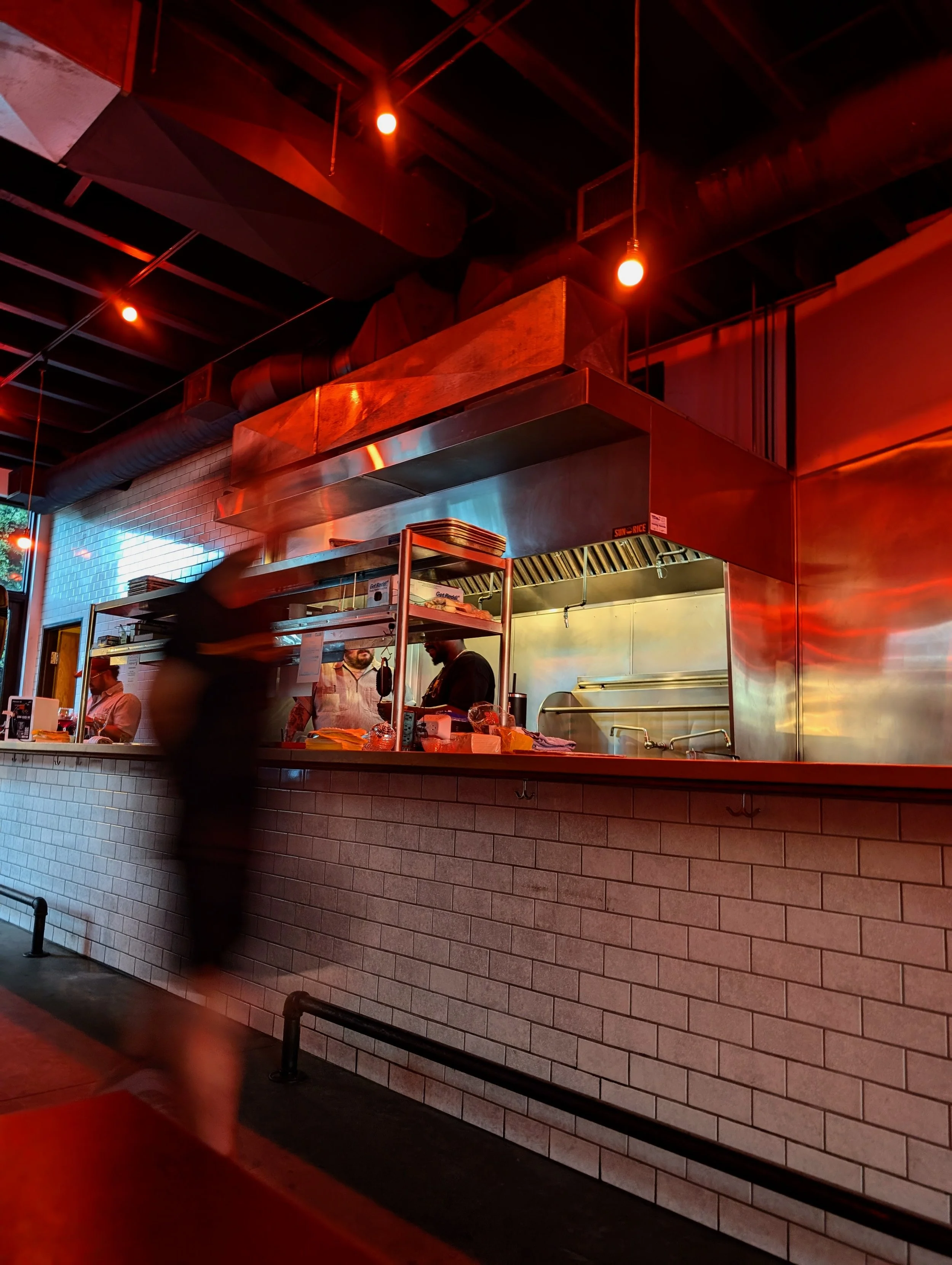Inside a modern restaurant kitchen with a brick counter and open window view showing chefs working with stainless steel equipment, illuminated by orange and red lighting.