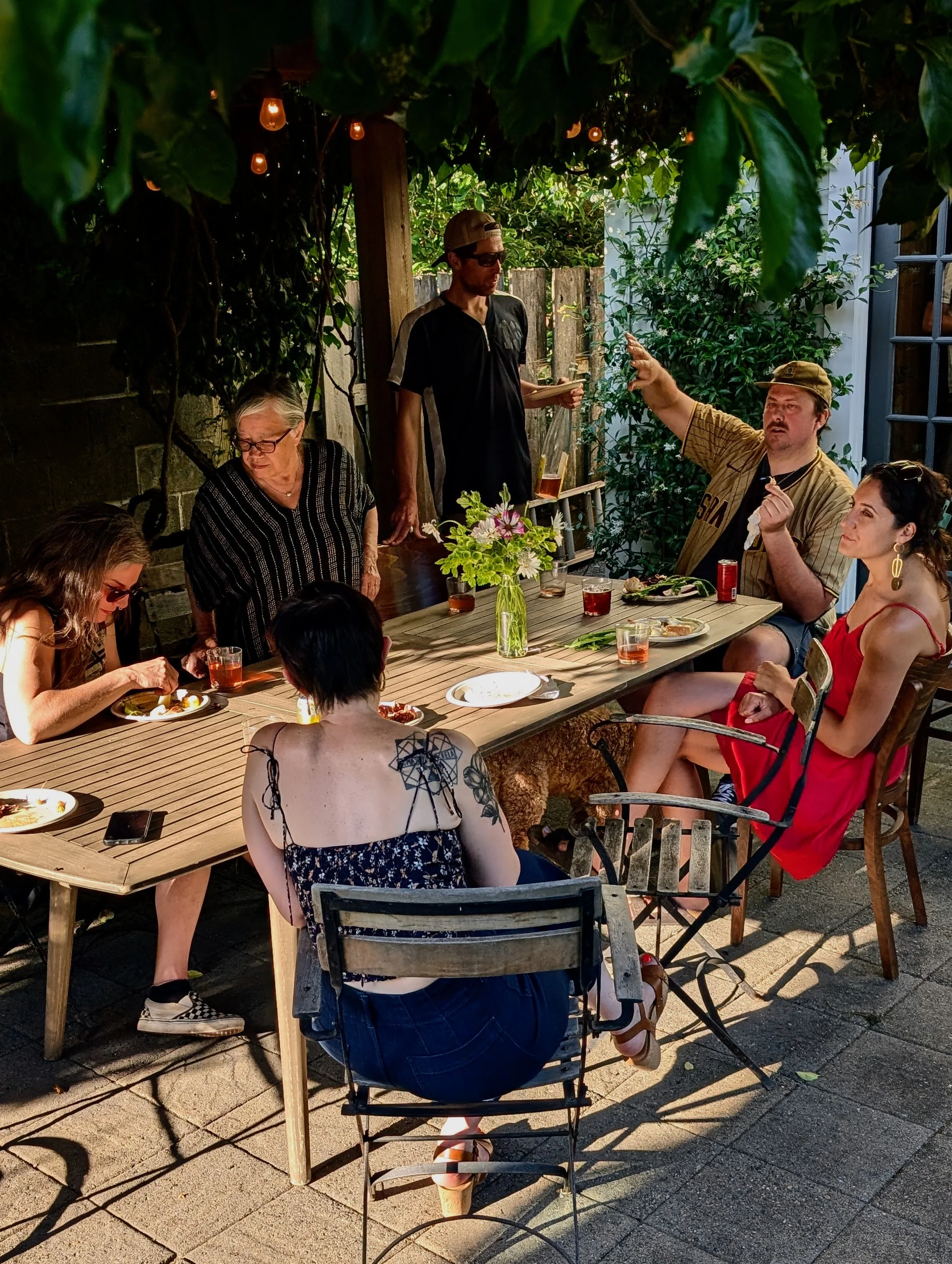 A group of people gathered around a wooden outdoor table, some eating, with a man in a baseball cap and sunglasses standing near the head of the table talking. There are drinks, a vase with flowers, and string lights overhead in a backyard setting.