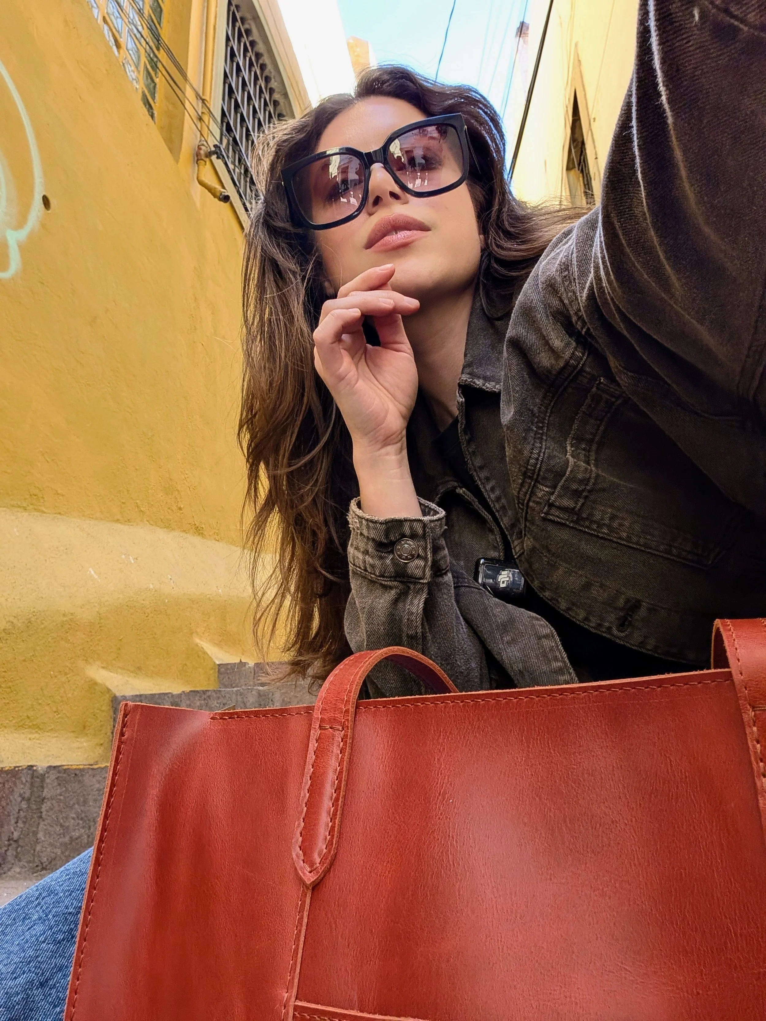A woman wearing large sunglasses and a denim jacket takes a selfie outdoors against a yellow wall with graffiti, with a large red purse in the foreground.