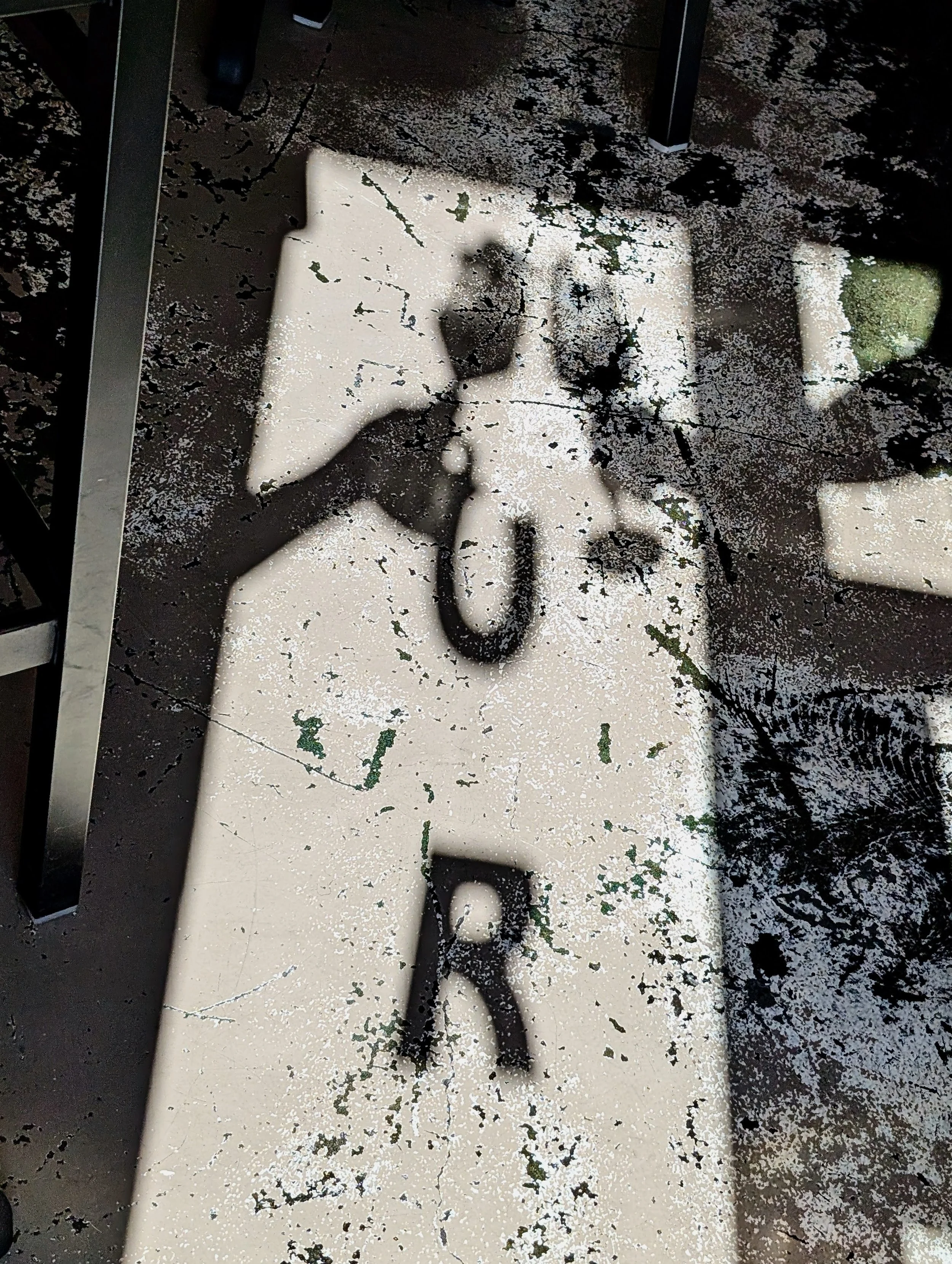 Shadow of a person taking a photo, reflected on a worn, cracked table with white paint, black paint splatters, and green patches.