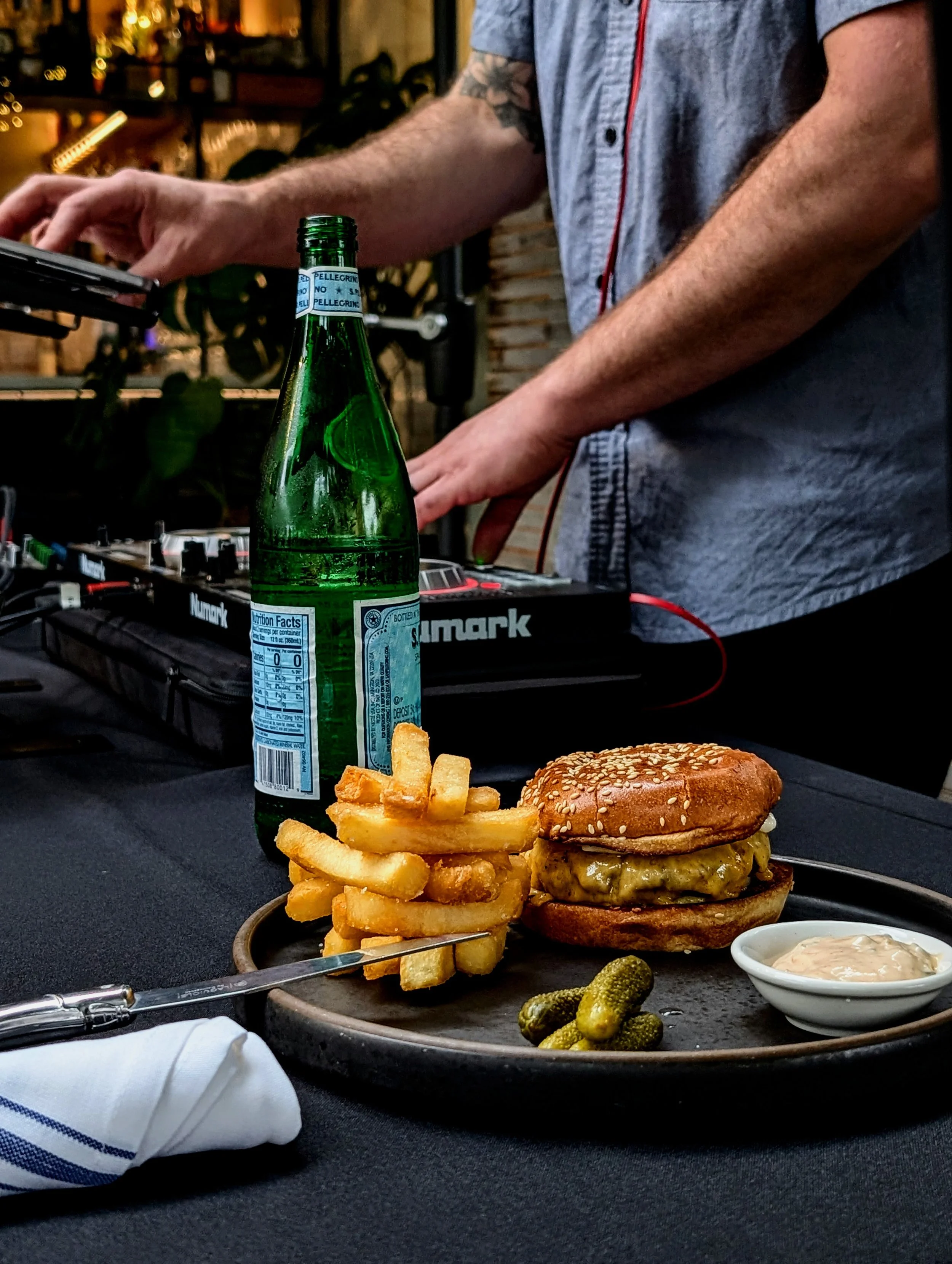 Close-up of a black plate with a cheeseburger, French fries, pickles, and a side of dipping sauce on a black table. A green glass bottle of sparkling water is partially in view. In the background, a DJ is working on equipment, wearing a gray short-sl