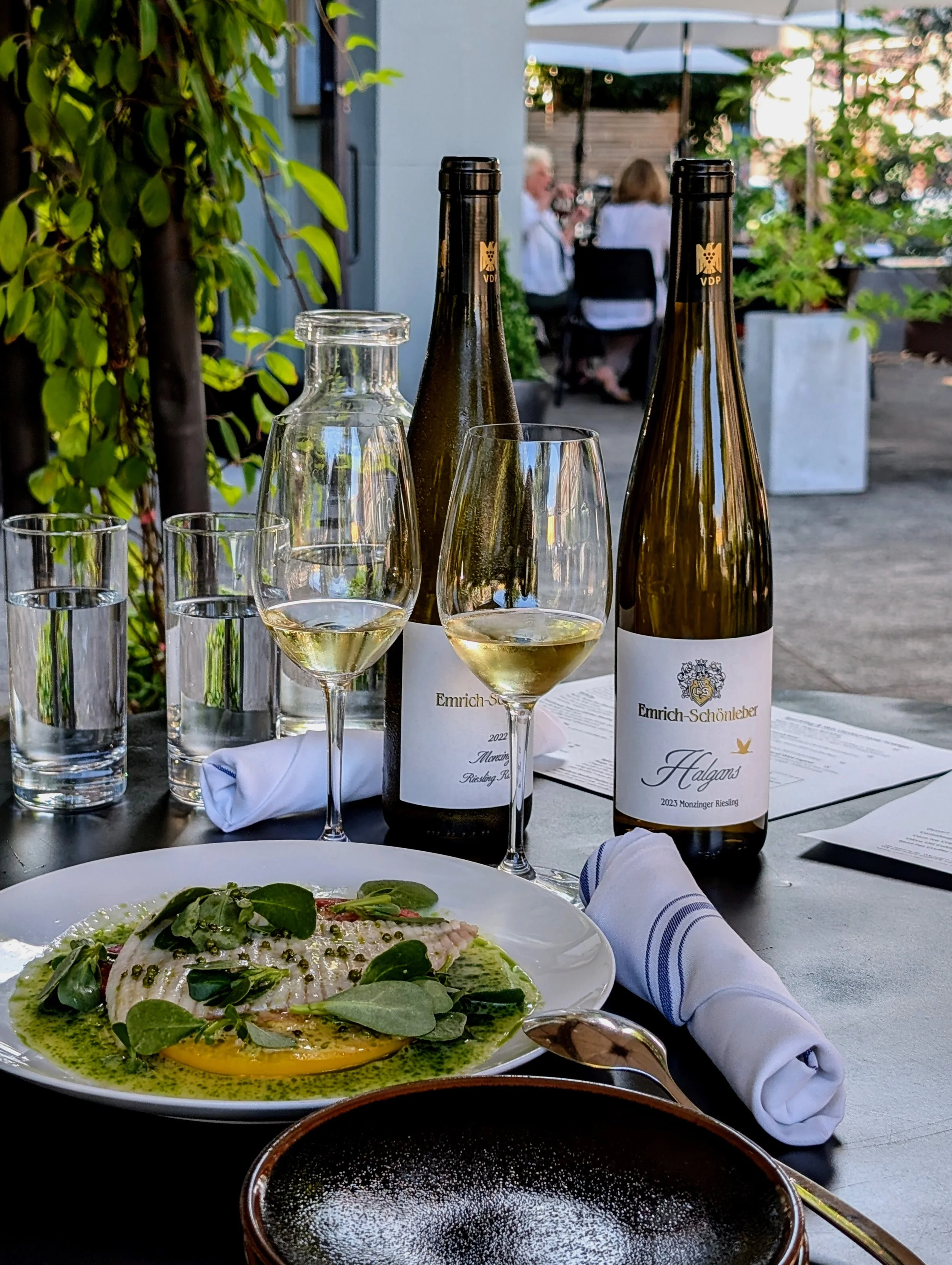 A table set for dining outdoors with two bottles of white wine, two wine glasses filled with white wine, and a plate of fish with greens and sauce. There are also three glasses of water and rolled napkins on the table.