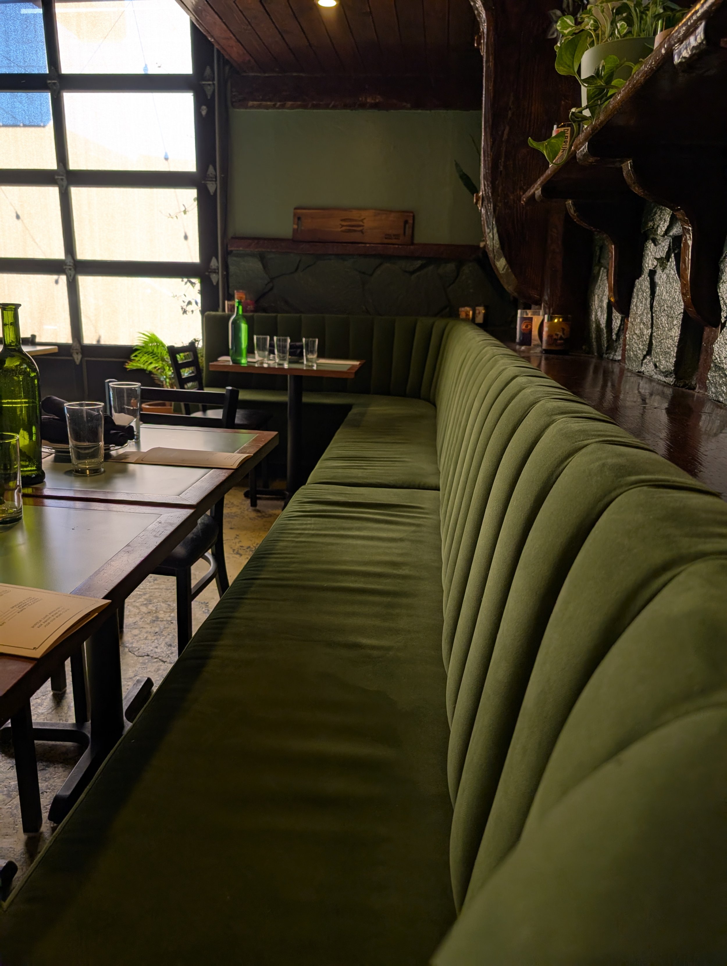Interior of a cozy restaurant with a long green velvet banquette along a wooden wall, tables set with water bottles, glasses, and menus, and large windows allowing natural light to fill the space.