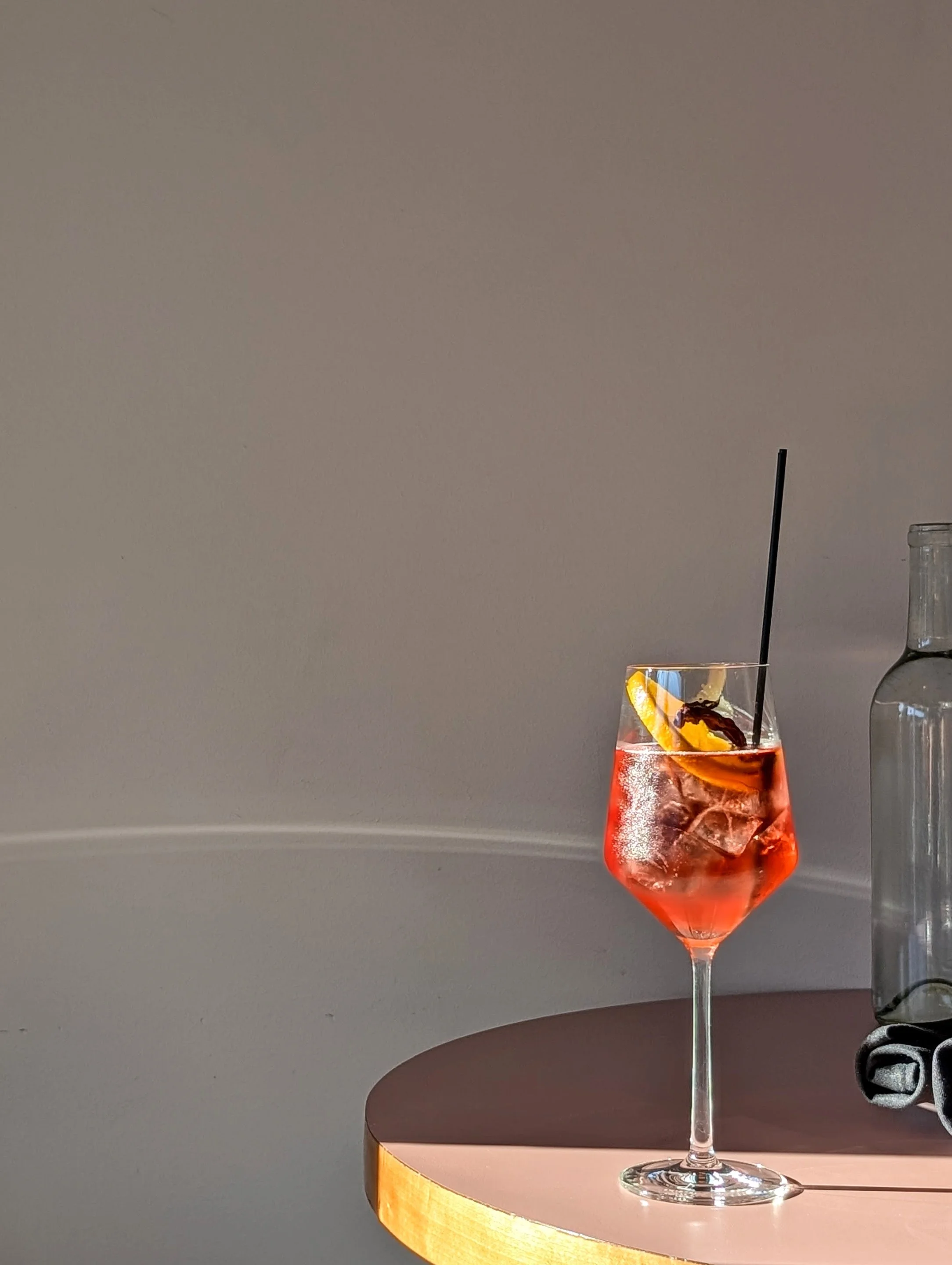 A pink cocktail with ice, garnished with an orange slice and a cherry, served in a tall, elegant glass with a black straw on a pink round table. A clear glass bottle is partially visible to the right.