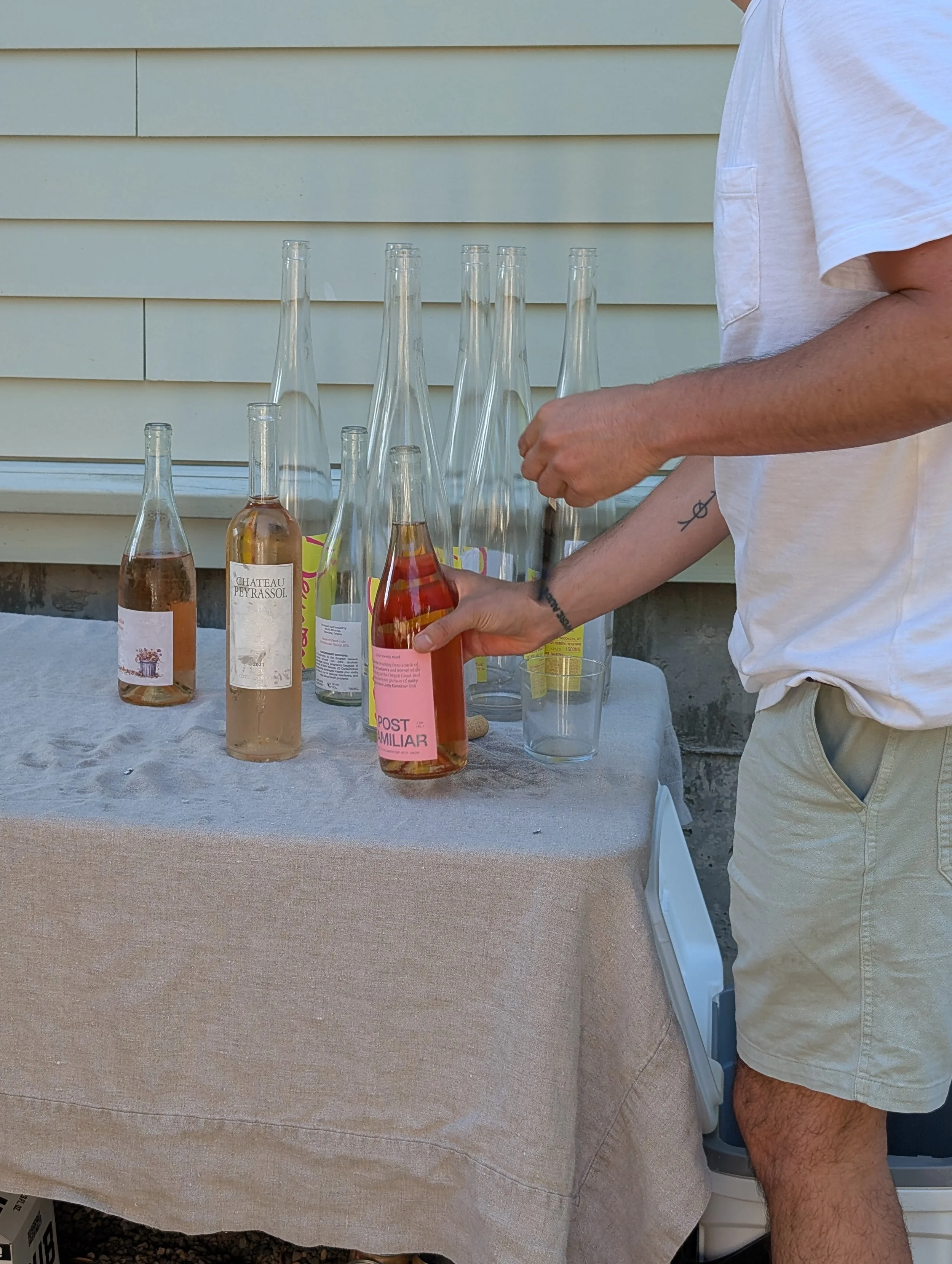 Person wearing a white T-shirt and beige shorts arranging empty and partially filled glass bottles on a table covered with a beige cloth outdoors against a house wall.
