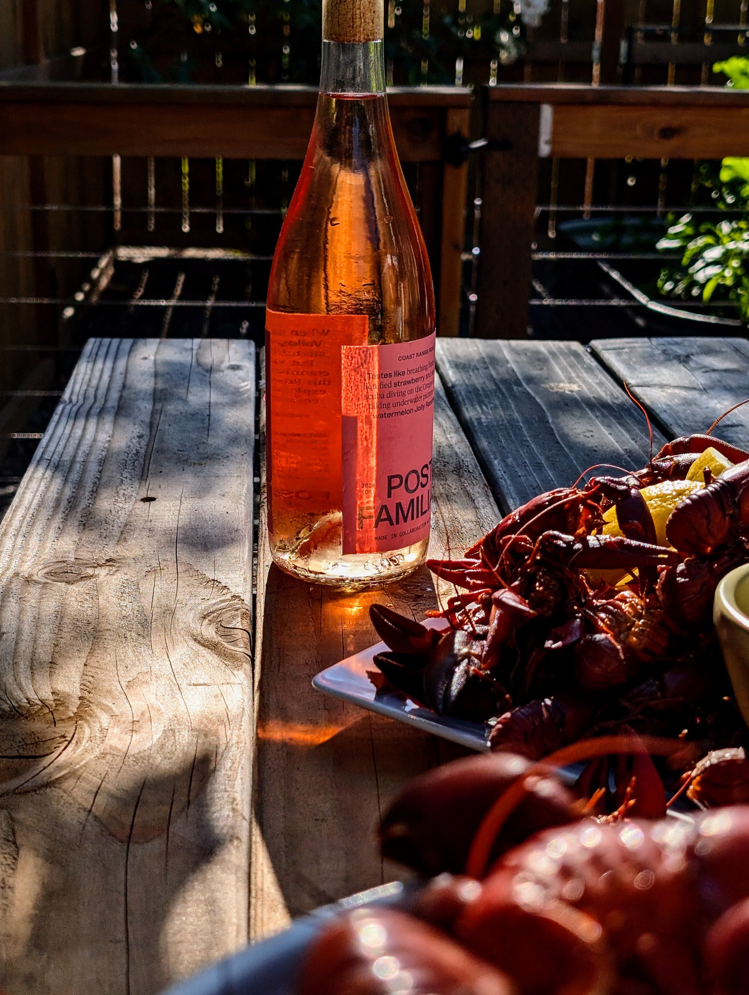 A bottle of rosé wine on a wooden table next to a plate of cooked crawfish and lemon wedges, with a background of a wooden fence and greenery.