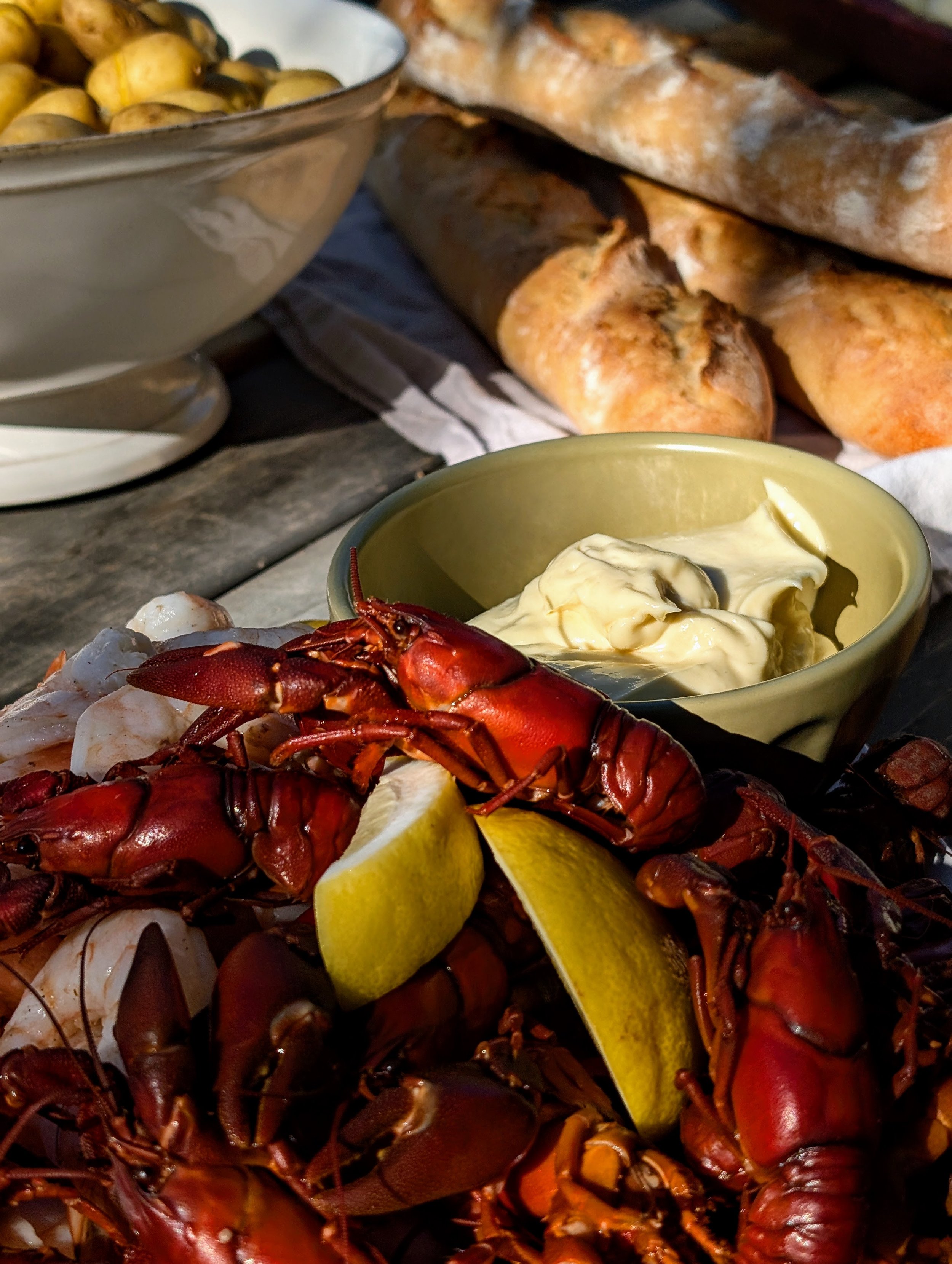 Lobster cooked with lemon wedges, served with a bowl of mayonnaise or aioli, with bread rolls and a bowl of small potatoes in the background.