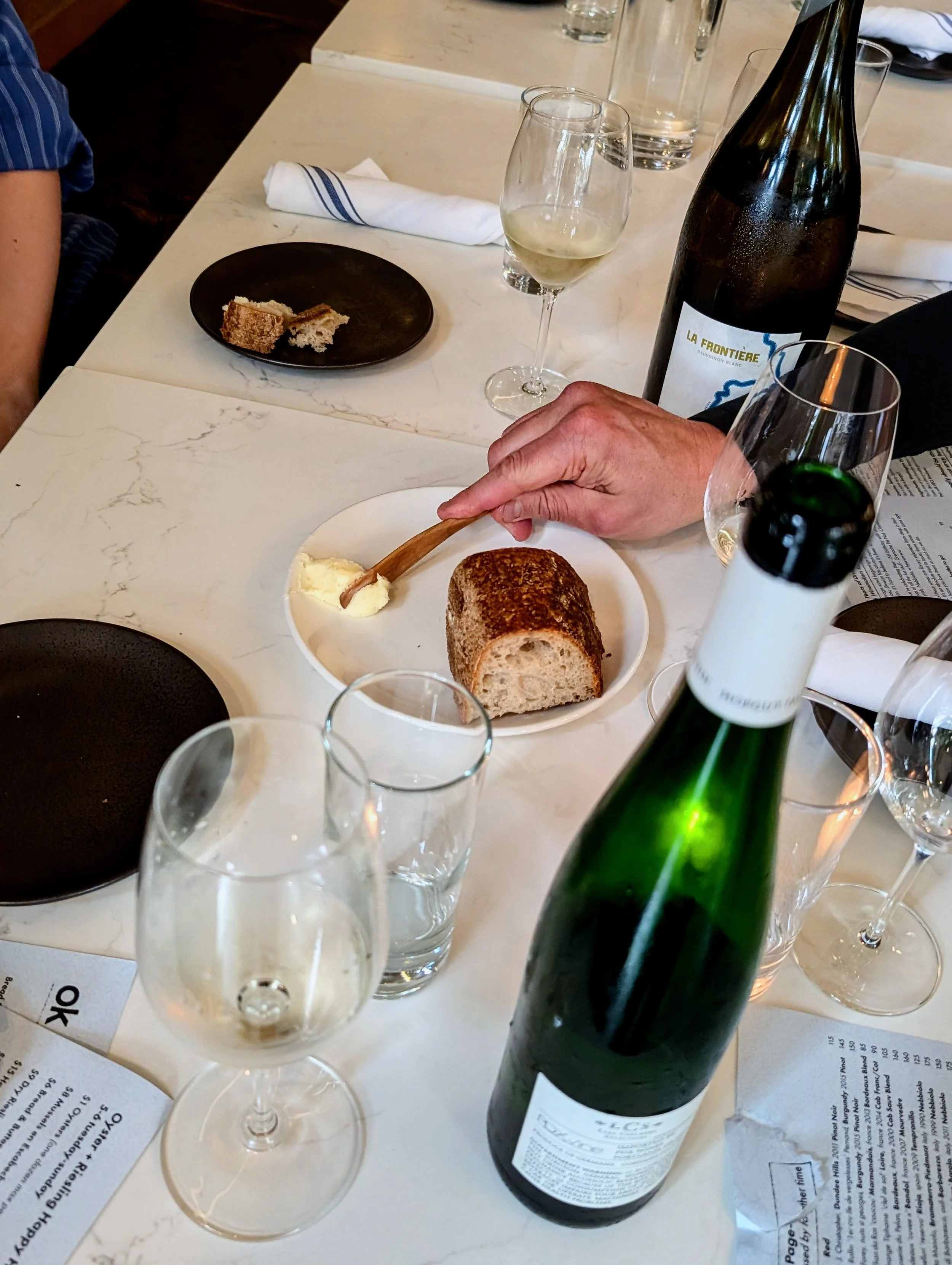 A table at a restaurant with various glasses of white wine, a bottle of wine, a piece of bread on a plate, and some tablesetting items including napkins and utensils.