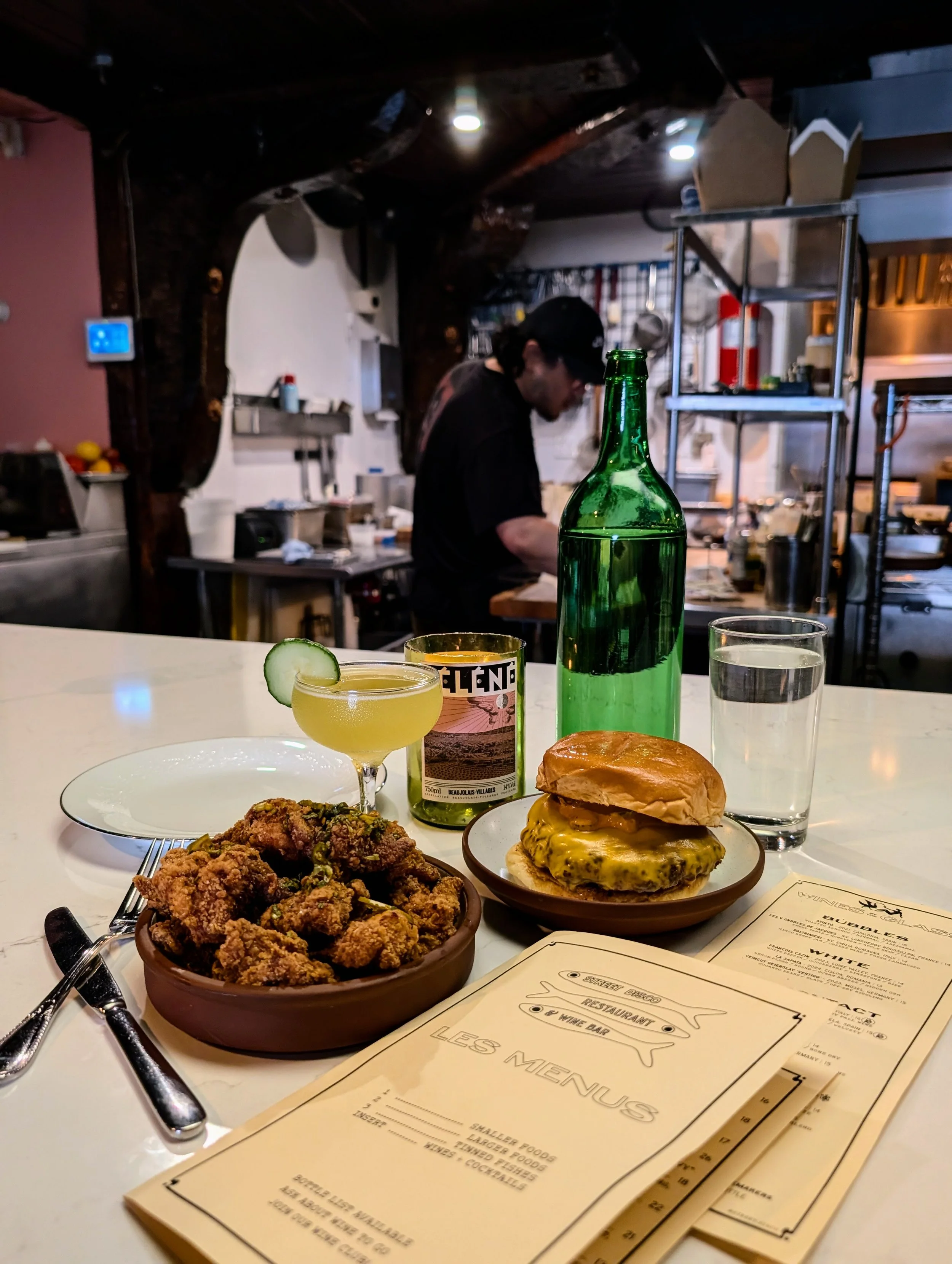 A table in a restaurant with a plate of fried chicken, a cheeseburger with a bun on top, a cocktail garnished with a cucumber slice, a green glass bottle, a glass of water, and menus, with a chef working in the background.