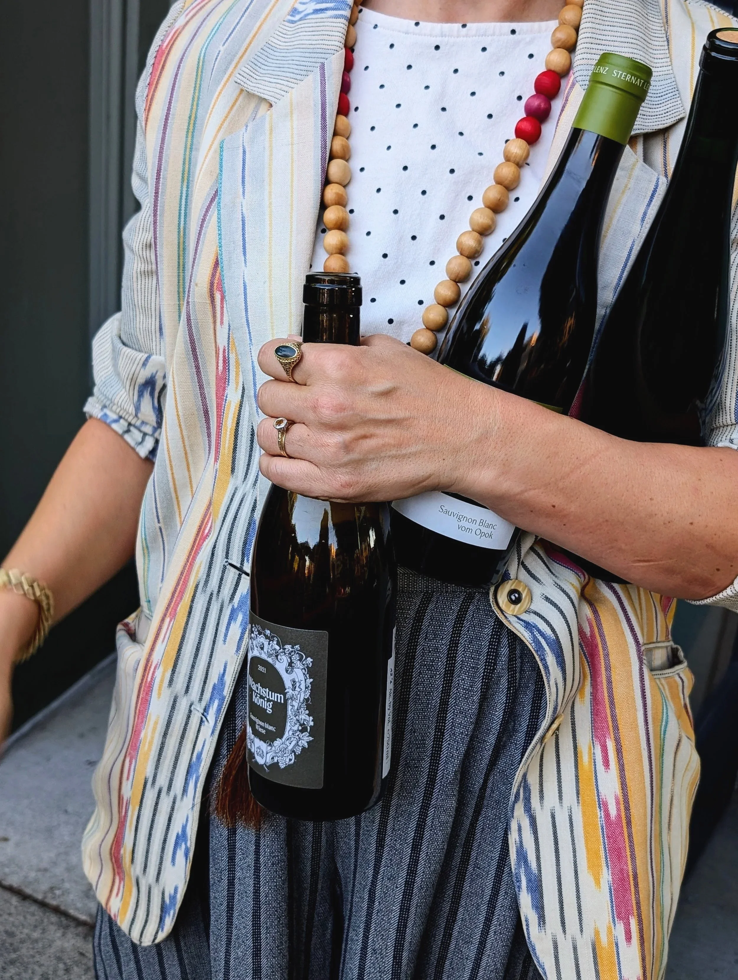 Person holding two bottles of wine, wearing a striped blazer, a polka dot shirt, and layered bead necklaces.