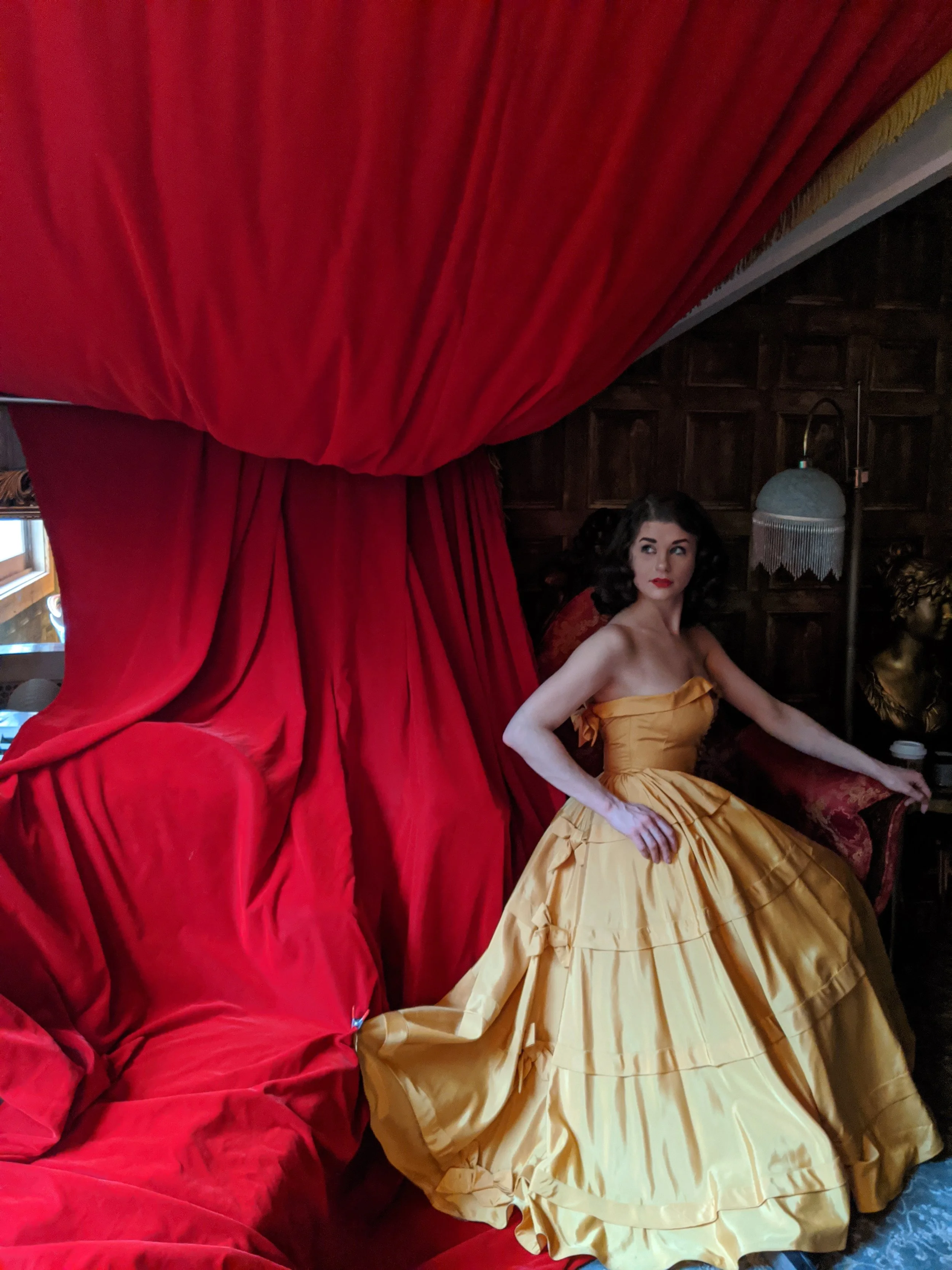A woman in a vintage yellow ball gown sitting on a red velvet chair in front of red curtains, in a dimly lit room with wooden walls and a vintage lamp.