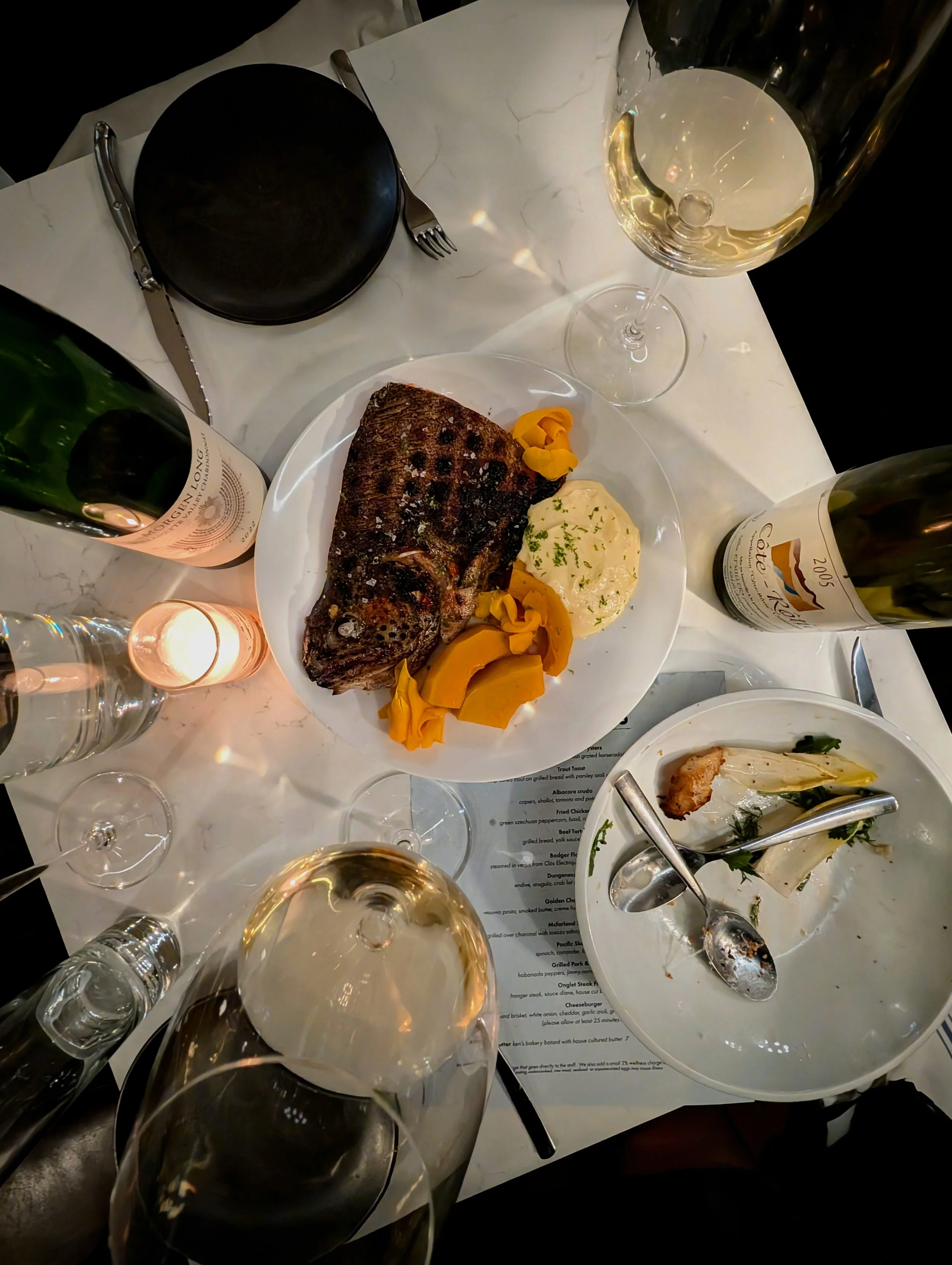 A table set for a meal with plates of grilled meat, mashed potatoes, vegetables, and wine glasses filled with white wine, along with a bottle of wine, water, and a candle.