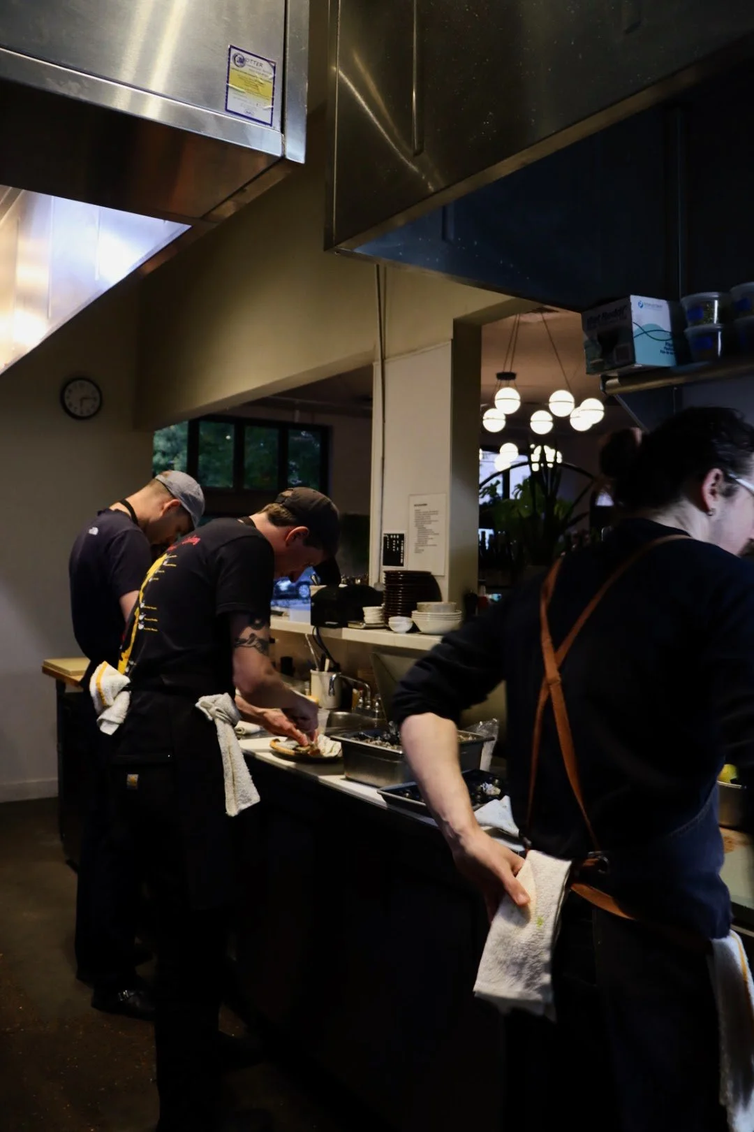 Three chefs preparing food in a busy commercial kitchen with stainless steel appliances, bowls, and kitchen tools on the counter, and decorative lighting in the background.