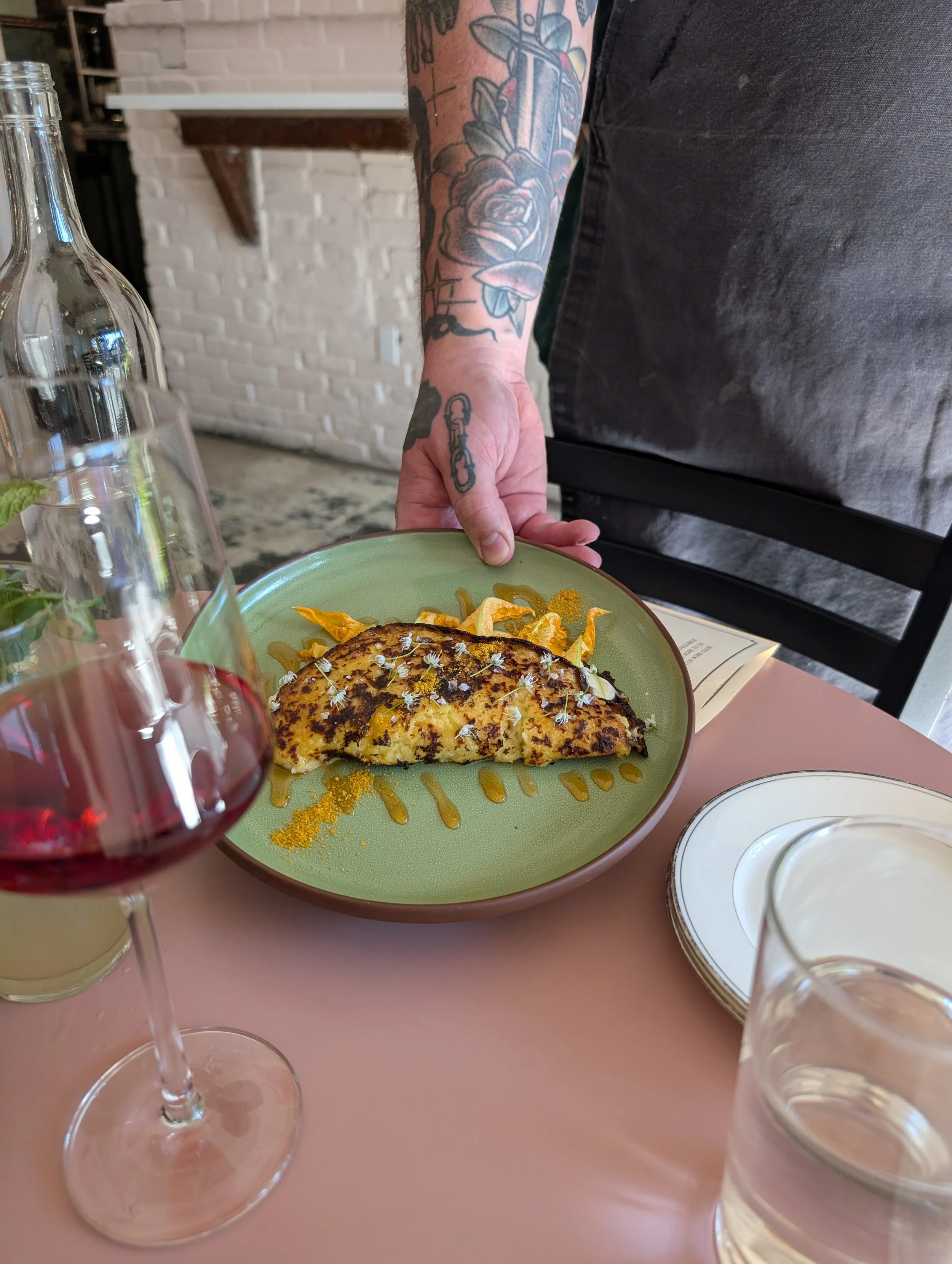 Person with tattoos holding a plate with a grilled cheese sandwich garnished with herbs and drizzled with sauce, on a pink table with wine and water glasses.