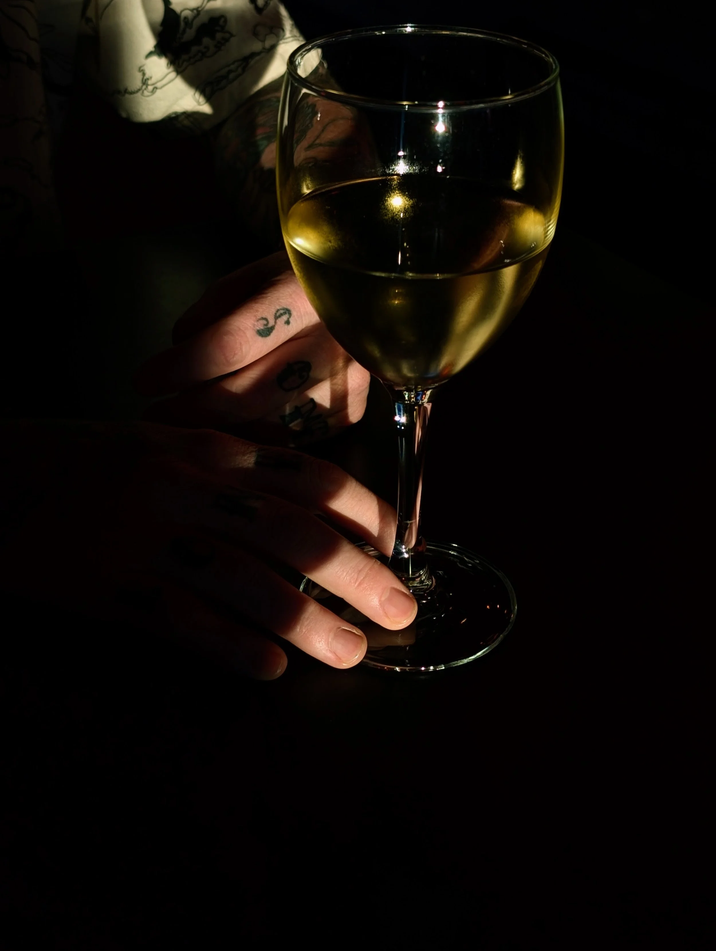 Hand holding a glass of white wine, with tattoos visible on fingers, in a dimly lit setting.