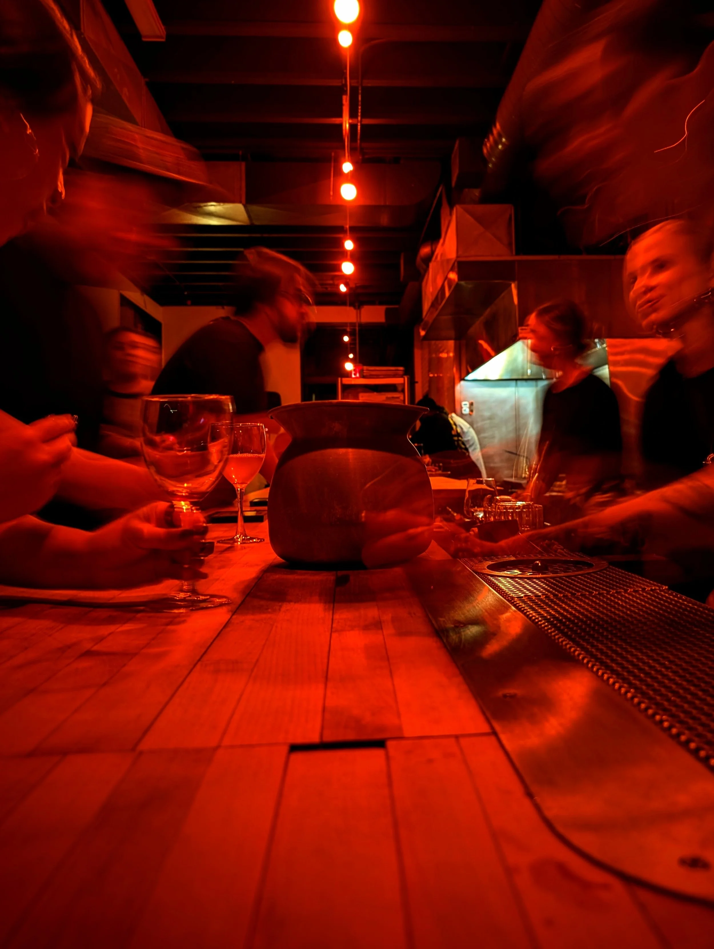 Group of people sitting at a bar illuminated by red lighting, with blurred motion. Glasses and a vase are on the wooden bar counter.