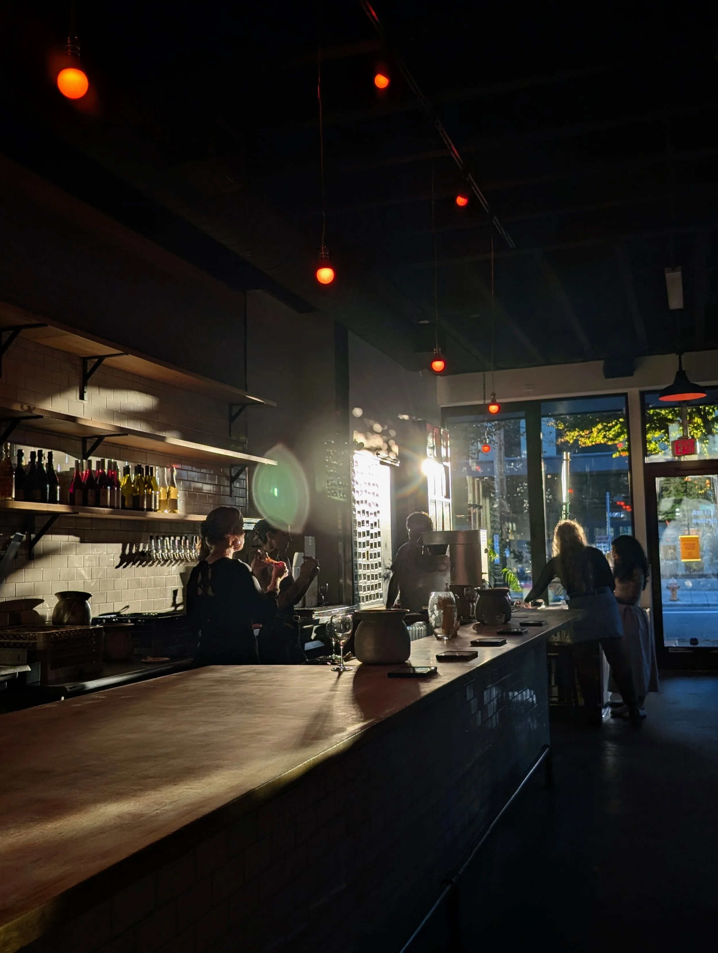 Indoor scene in a dimly lit bar or cafe with hanging red lights, a wooden countertop, bottles on shelves, and people near the window enjoying the sunset.