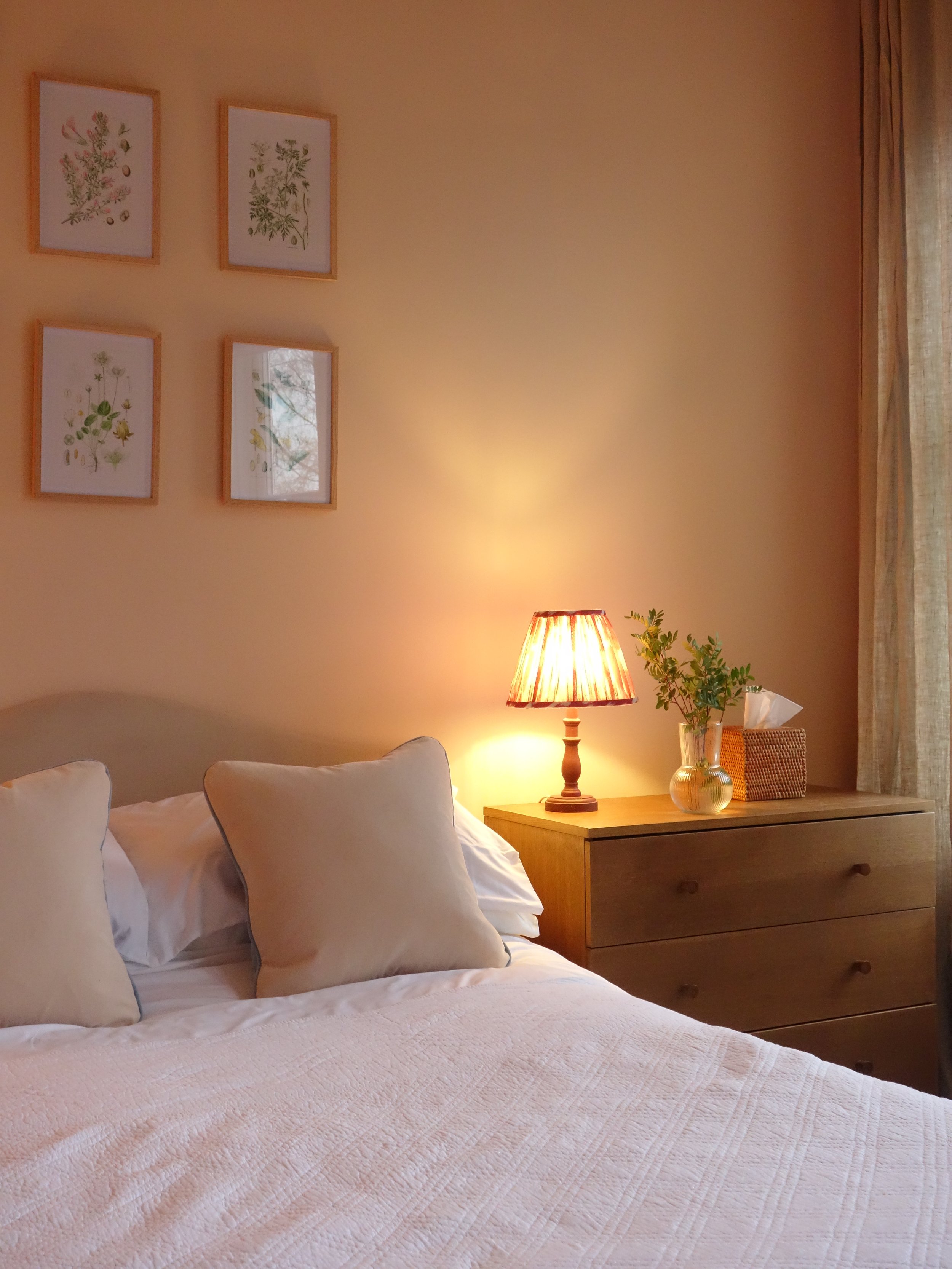 A cozy bedroom with a bed covered in a textured white quilt, four pillows, a wooden nightstand with a lamp emitting warm light, a potted plant, framed botanical artwork on the wall, and a curtain on the window.