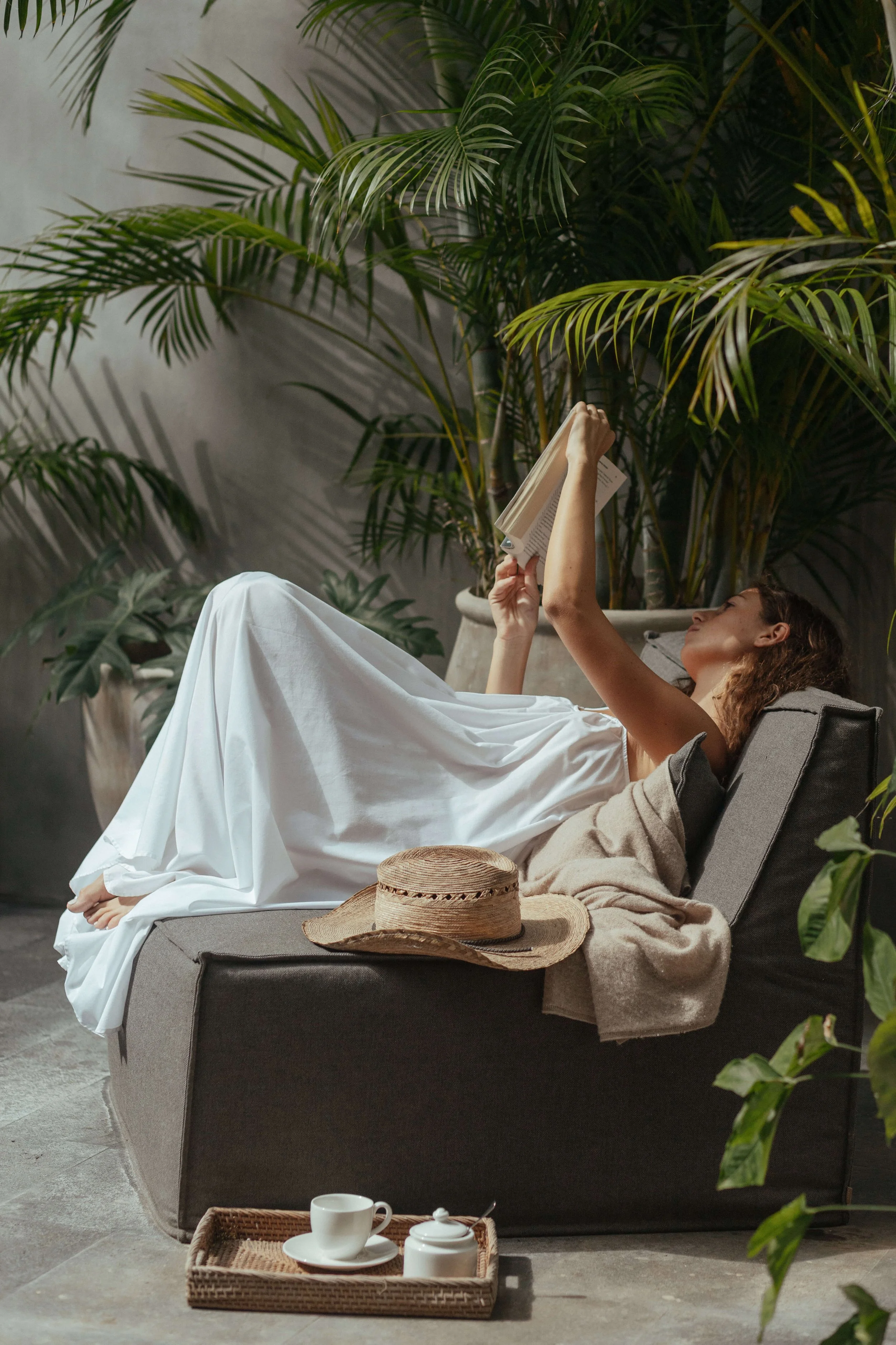 Sunlit modern room with beige textured walls, large decorative mirror, wooden chair, potted plants in large stone pots, exterior view through glass door, and minimal decor.