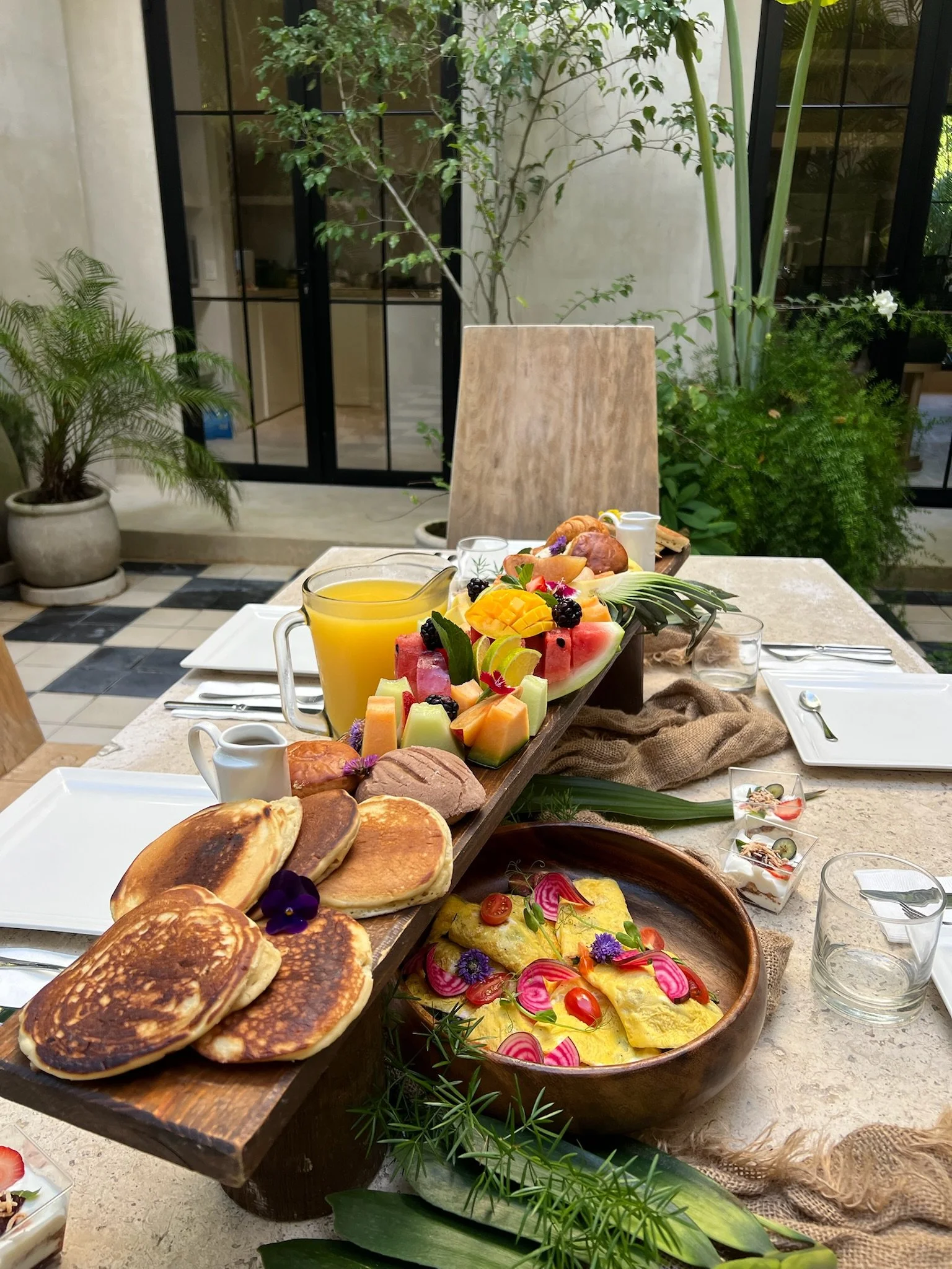 A breakfast table with pancakes, fresh fruit, juice, and small desserts held on a bamboo tray with wine glasses, plates, and utensils, set in a bright indoor space with plants and glass doors.