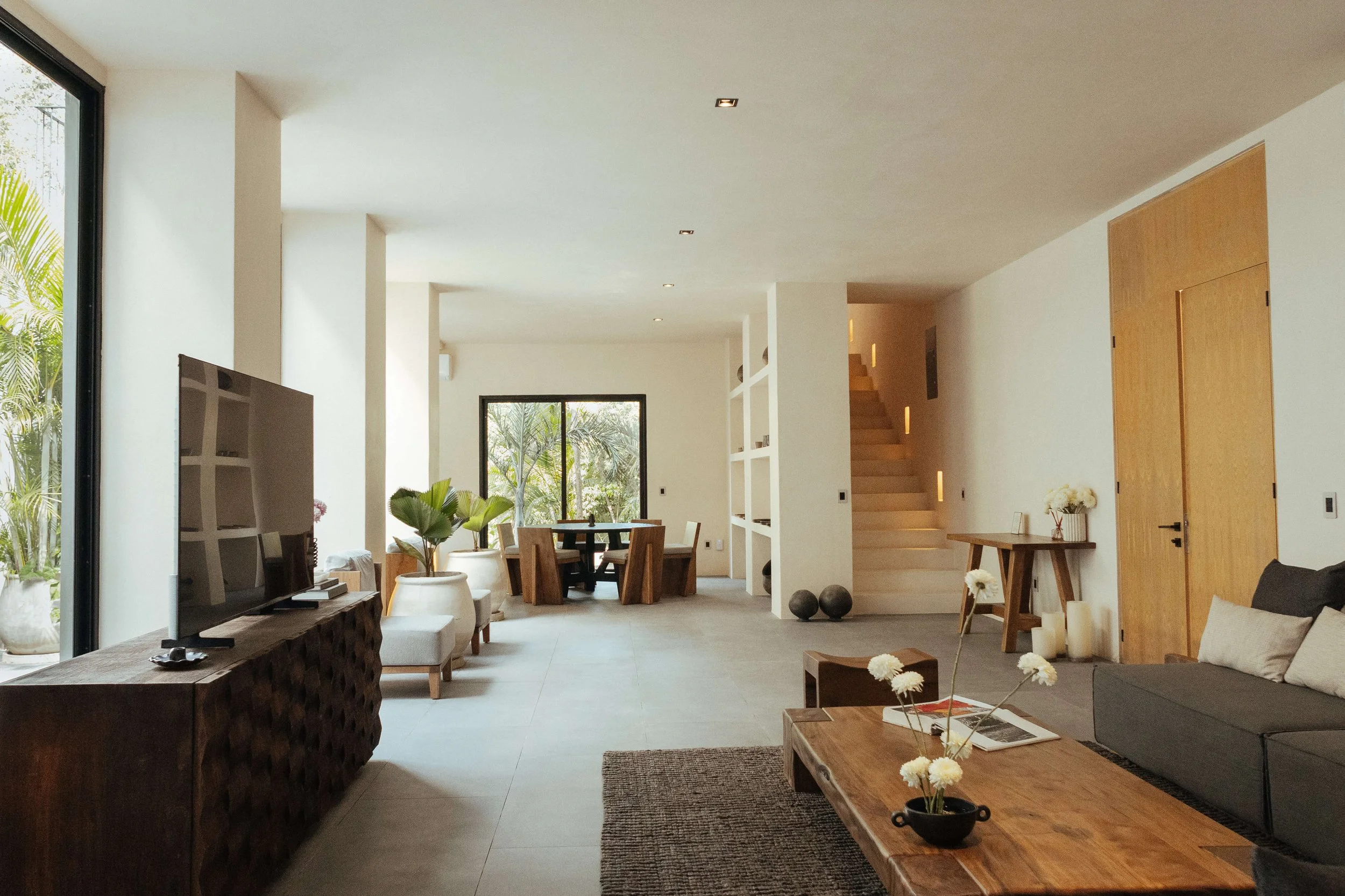 Modern living room with large gray sofa, two blue armchairs, wooden coffee table, potted plants, and built-in shelves, illuminated by natural light from large window.
