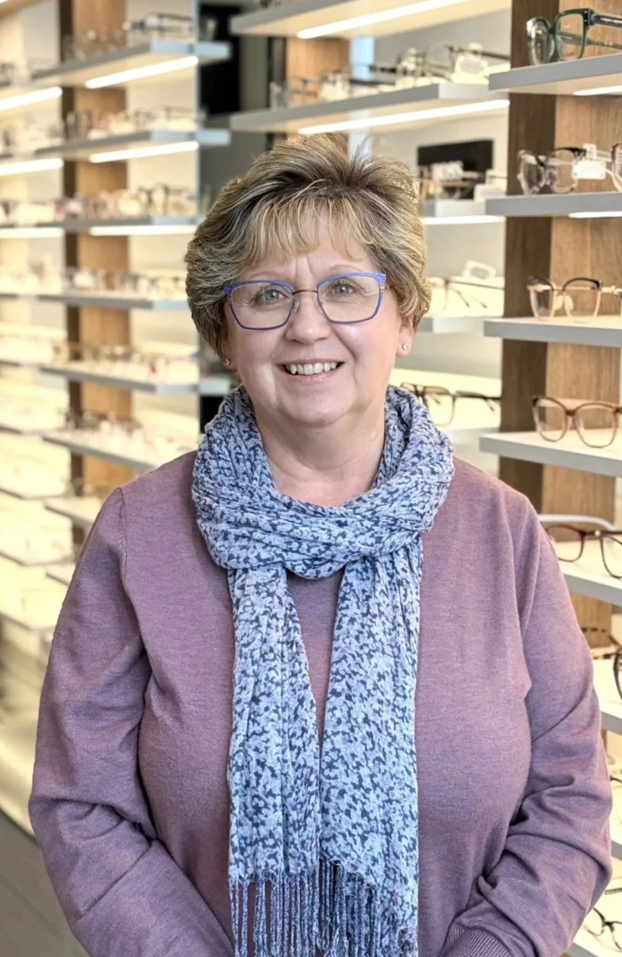 Woman with short, light brown hair and glasses smiling in an eyeglasses store, shelves with multiple pairs of eyeglasses in the background.