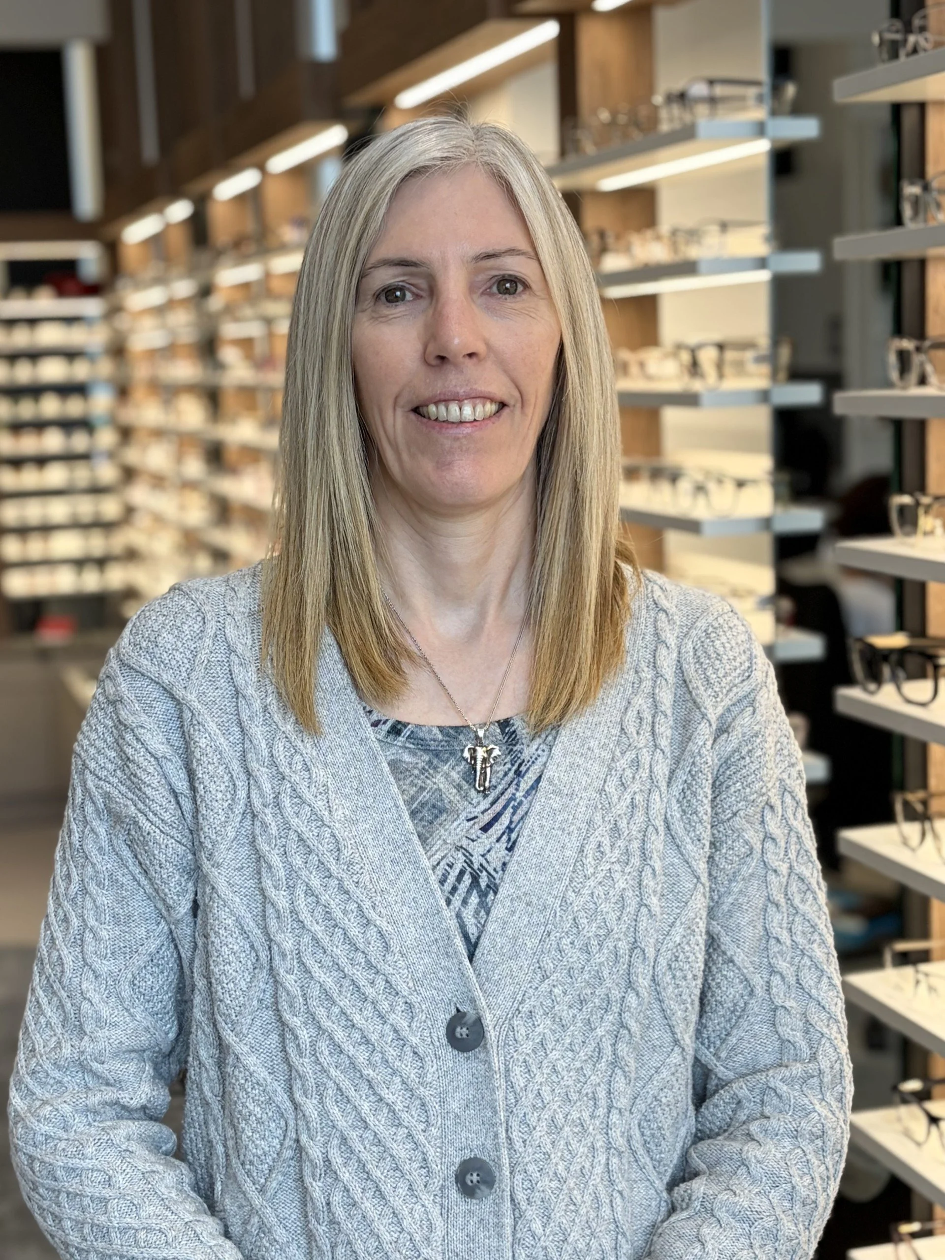 A woman with long, straight gray hair and fair skin stands in front of a display of eyeglasses, smiling at the camera. She is wearing a light gray, cable-knit cardigan over a patterned top and a silver necklace with a tree pendant.