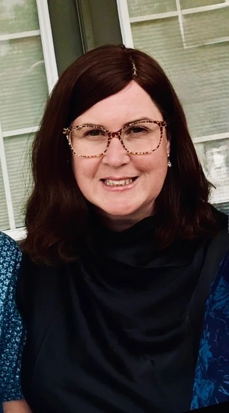 A woman with shoulder-length dark brown hair, wearing leopard print glasses and a black top, smiling in front of a window with blinds.