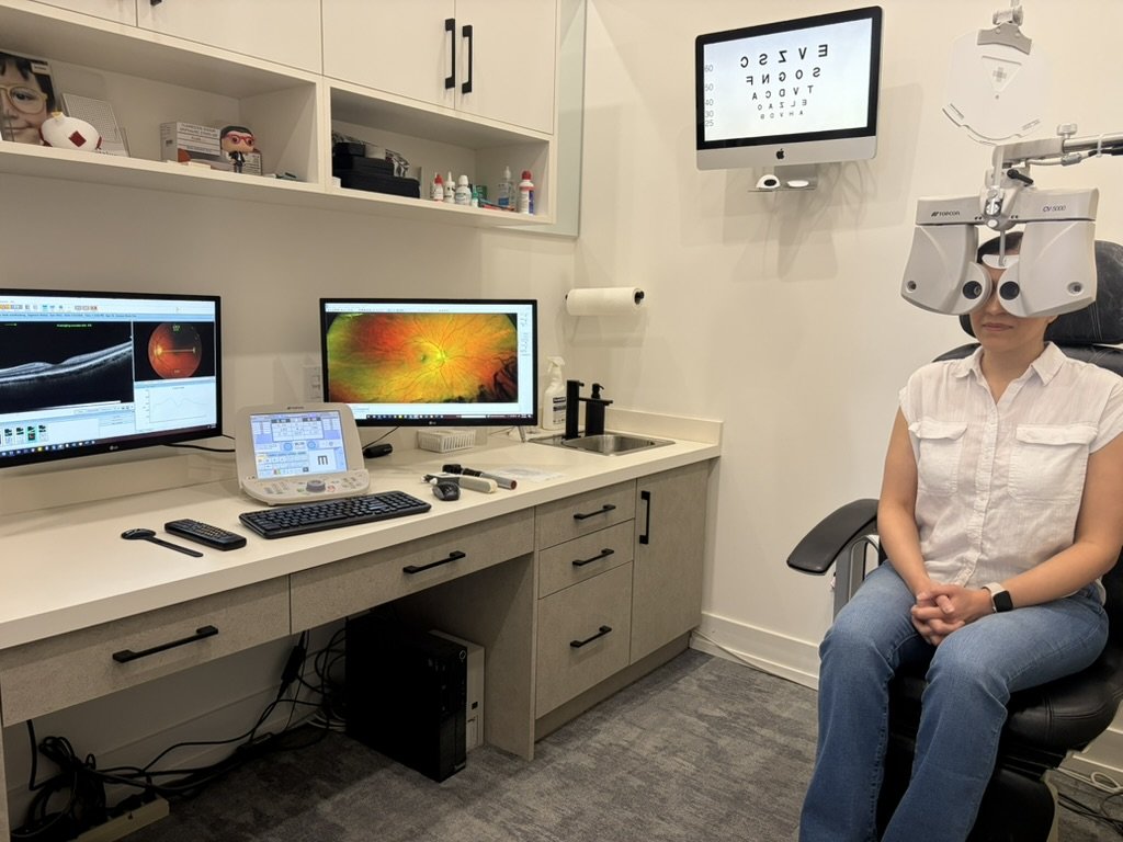 A woman in a white shirt and jeans sitting in an ophthalmologist's examination room while wearing a phoropter for eye examination. There are two computer monitors, one displaying an eye examination chart and the other showing an eye image. Additional ophthalmic equipment and supplies are on the countertop and shelving.