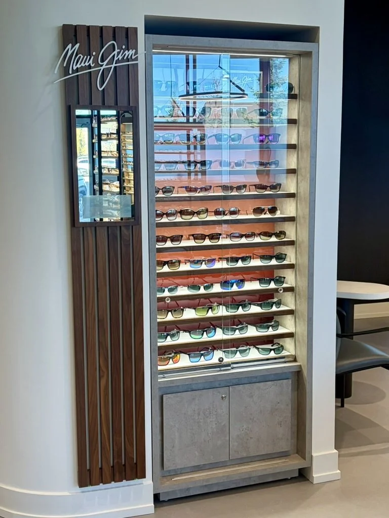 Display of Maui Jim sunglasses on a multi-shelf stand inside a store, with a mirror on the left side and a curve of a white wall, wood paneling, and a table with a chair on the right.