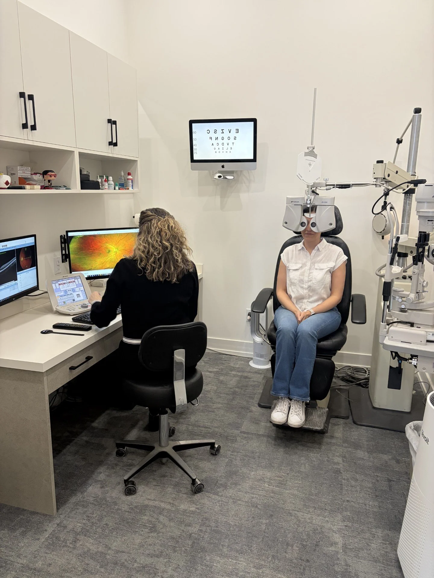Optometrist and patient in an eye exam room, with eye chart on wall and eye examination equipment.