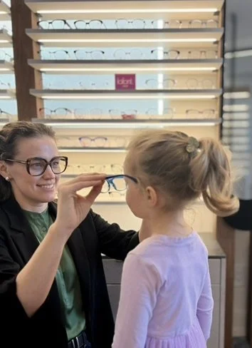 A woman helping a young girl try on glasses at an eyeglasses shop.