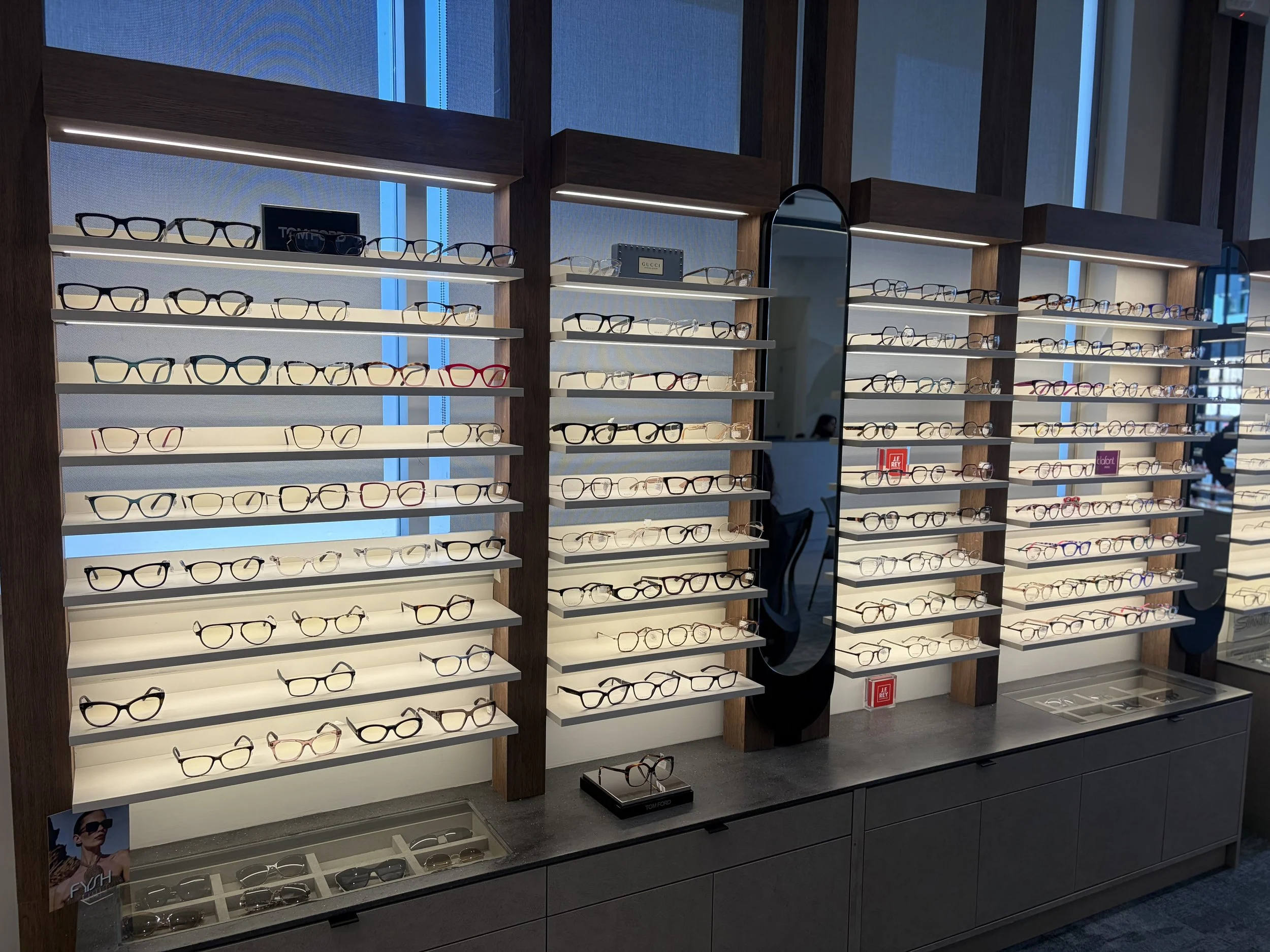 Display of multiple eyeglasses on white shelves with wooden framing, reflecting in a mirror in an eyewear store.