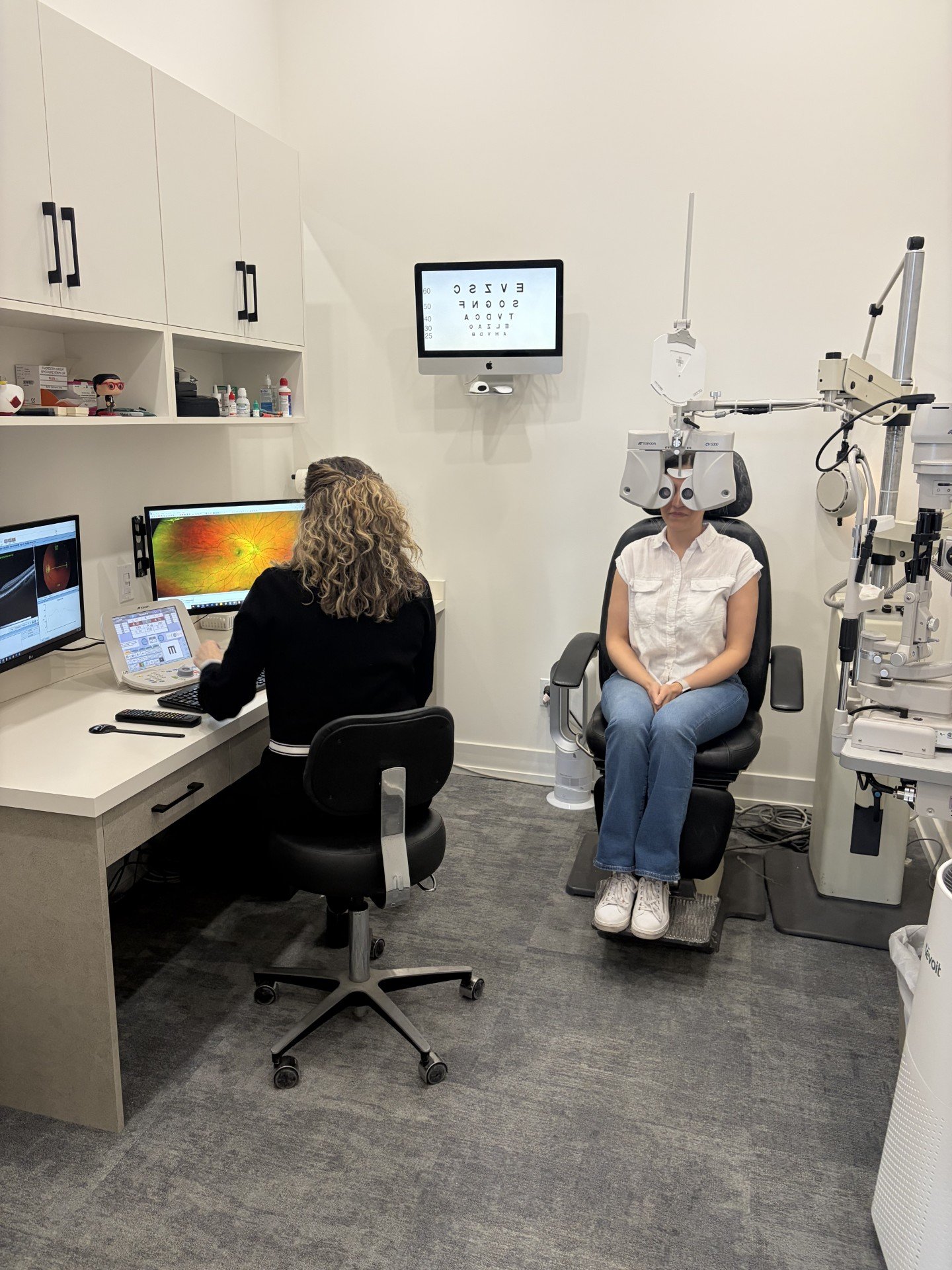 Patient sitting in an eye examination chair with specialized eye equipment, optometrist at computer, eye chart on screen.