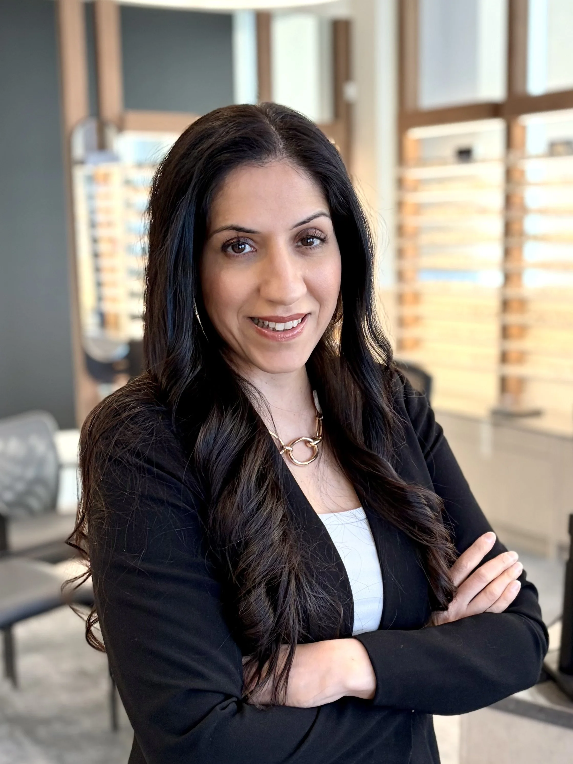 A woman with long dark hair, wearing a black blazer and jewelry, smiling with arms crossed in an office setting with natural light and modern decor.