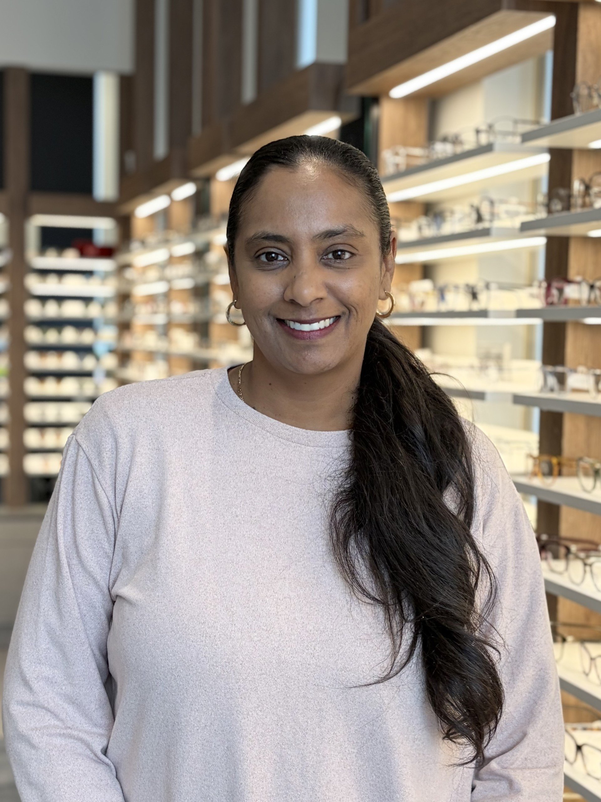 A woman with dark hair in a ponytail and wearing hoop earrings off a white shirt, smiling in an eyewear or accessory store with shelves of glasses in the background.