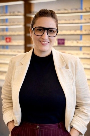 A woman with brown hair in a bun, wearing glasses, a black top, a light-colored blazer, and maroon pants, standing in a store aisle with shelves in the background.