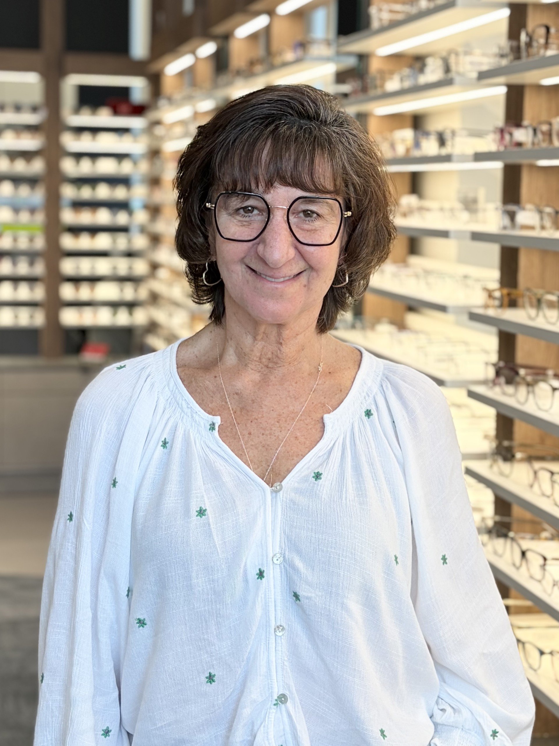 A woman with short brown hair and glasses smiling in an eyewear store with shelves of glasses in the background.