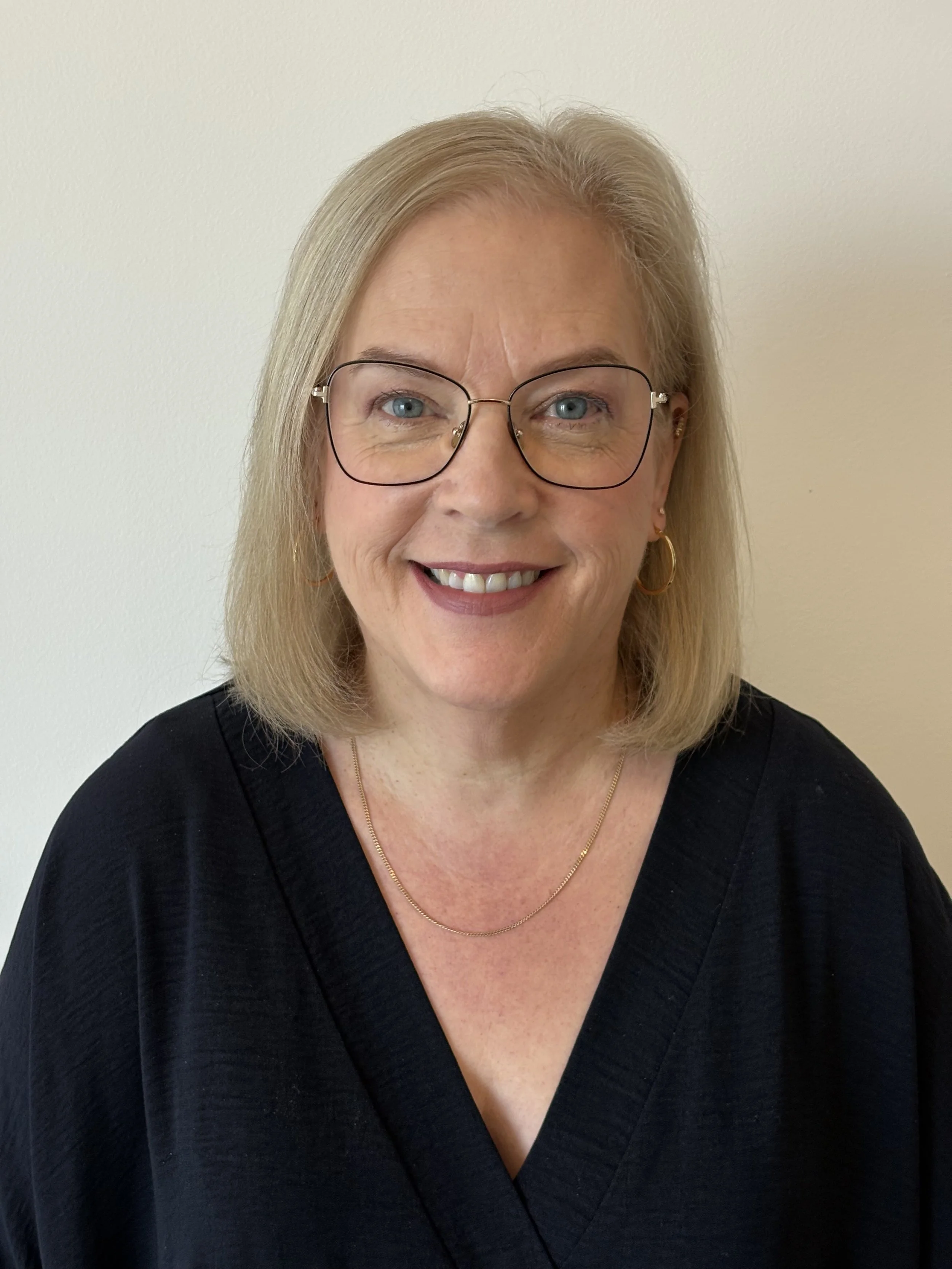 Middle-aged woman with shoulder-length blonde hair, wearing glasses, gold hoop earrings, a gold chain necklace, and a black top, smiling against a plain beige wall.