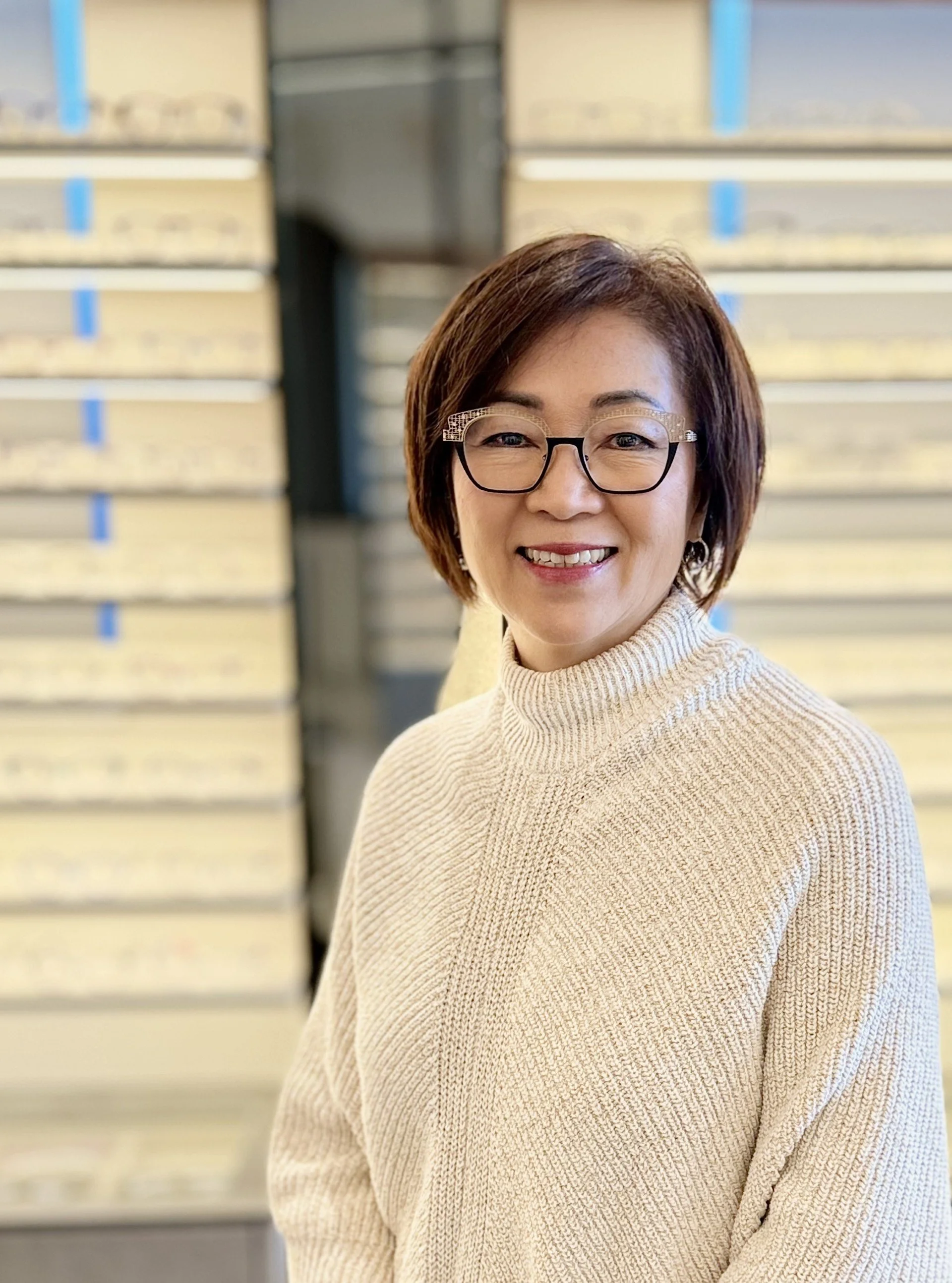 A woman with short brown hair and glasses smiling in front of a background of stacked boxes with blue labels.