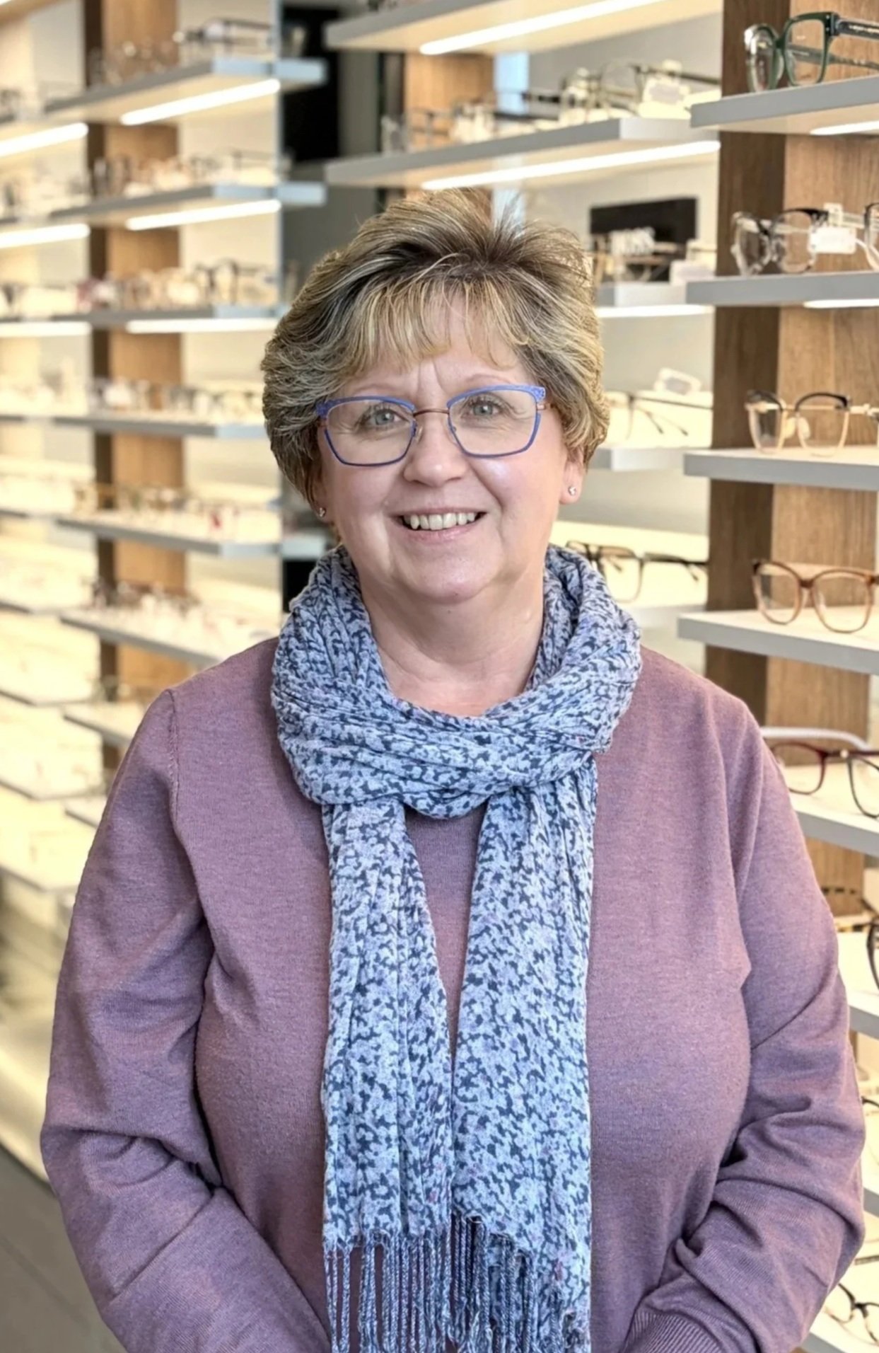 A woman with short blonde hair and glasses smiling inside an eyewear store, with shelves of eyeglasses in the background.