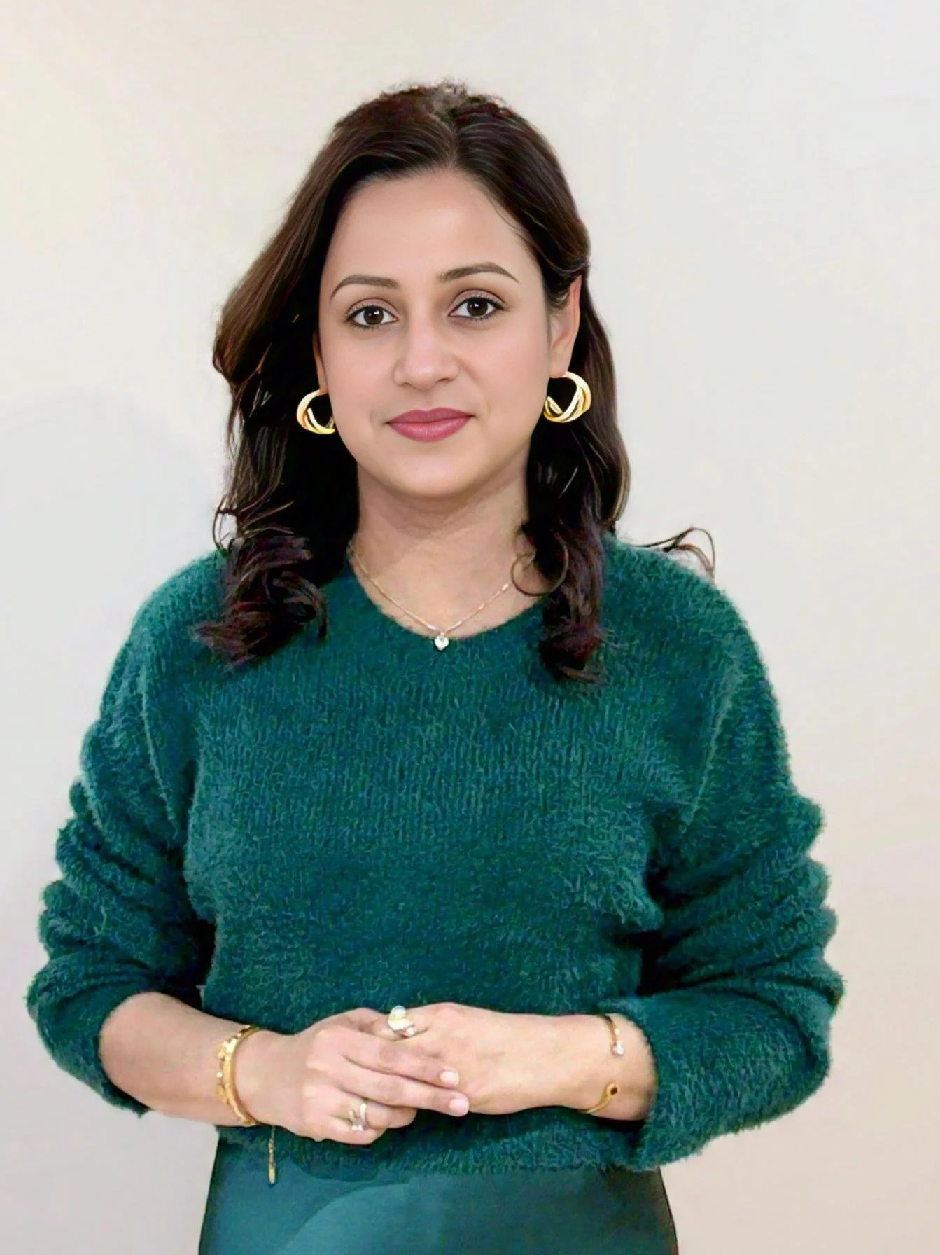 A woman with dark brown hair, wearing gold hoop earrings, a delicate necklace, and a yellow bracelet, dressed in a teal textured sweater, standing against a plain off-white background.