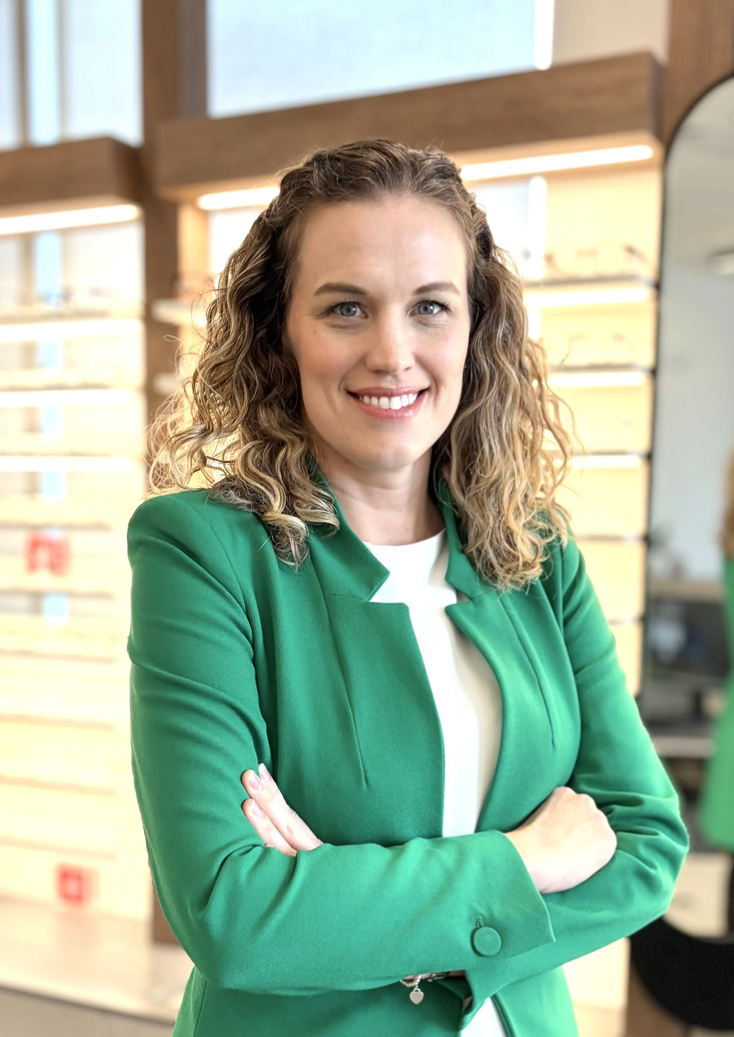 A woman with curly brown hair, blue eyes, and fair skin, smiling and crossing her arms, wearing a green blazer over a white top, in what appears to be an office environment.