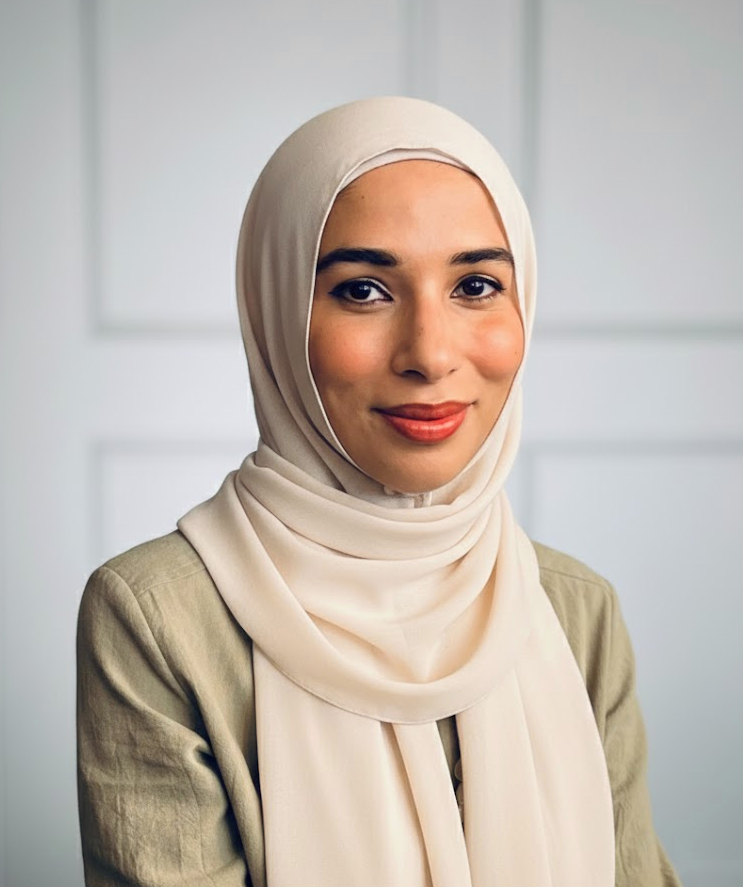 A woman wearing a beige hijab and a light-colored blazer, smiling in front of a plain light-colored background.