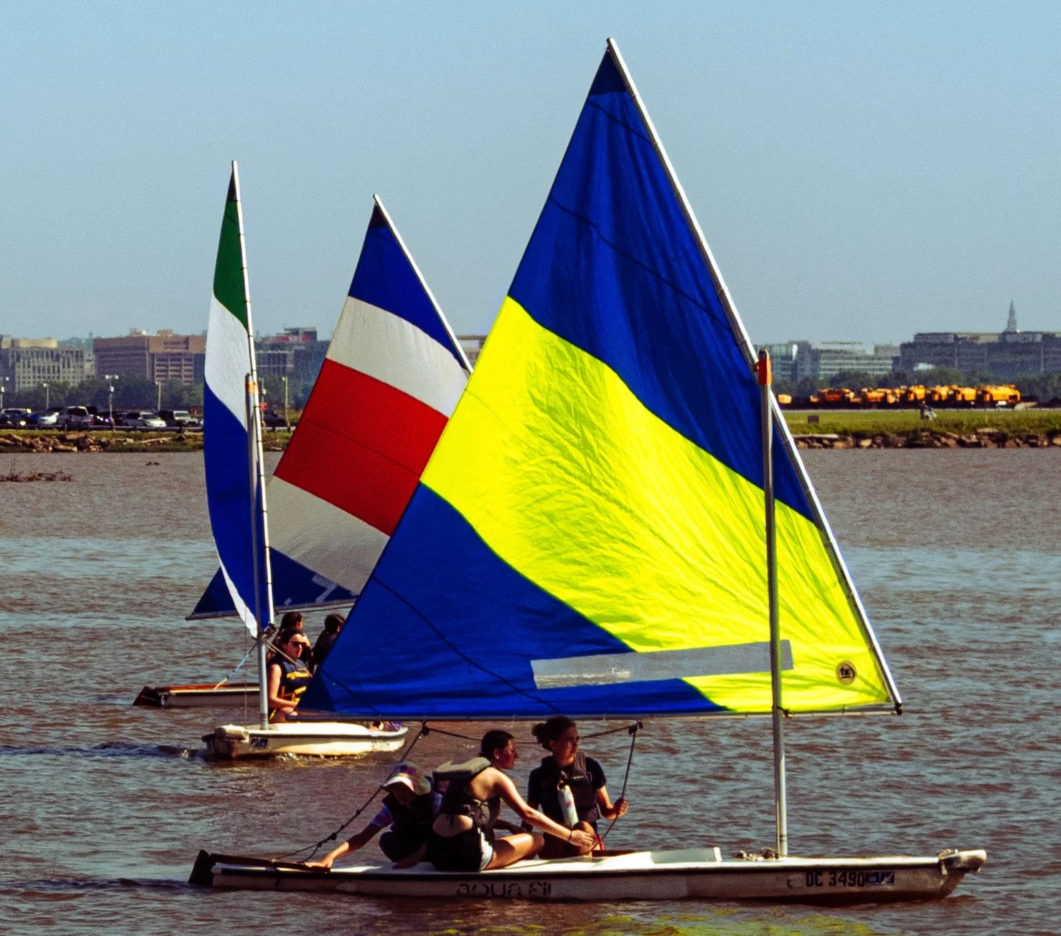 Young sailors navigate colorful dinghies on the Potomac River with the U.S. Capitol visible in the distance.