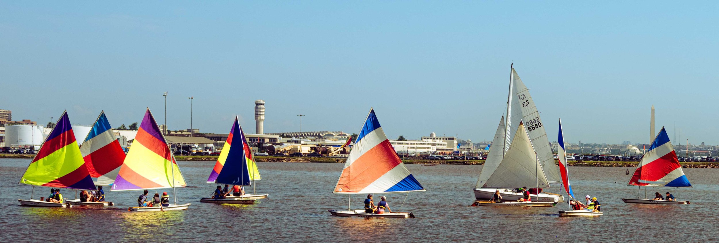 A colorful training sail glides across the river with the nation's capital visible in the distance.