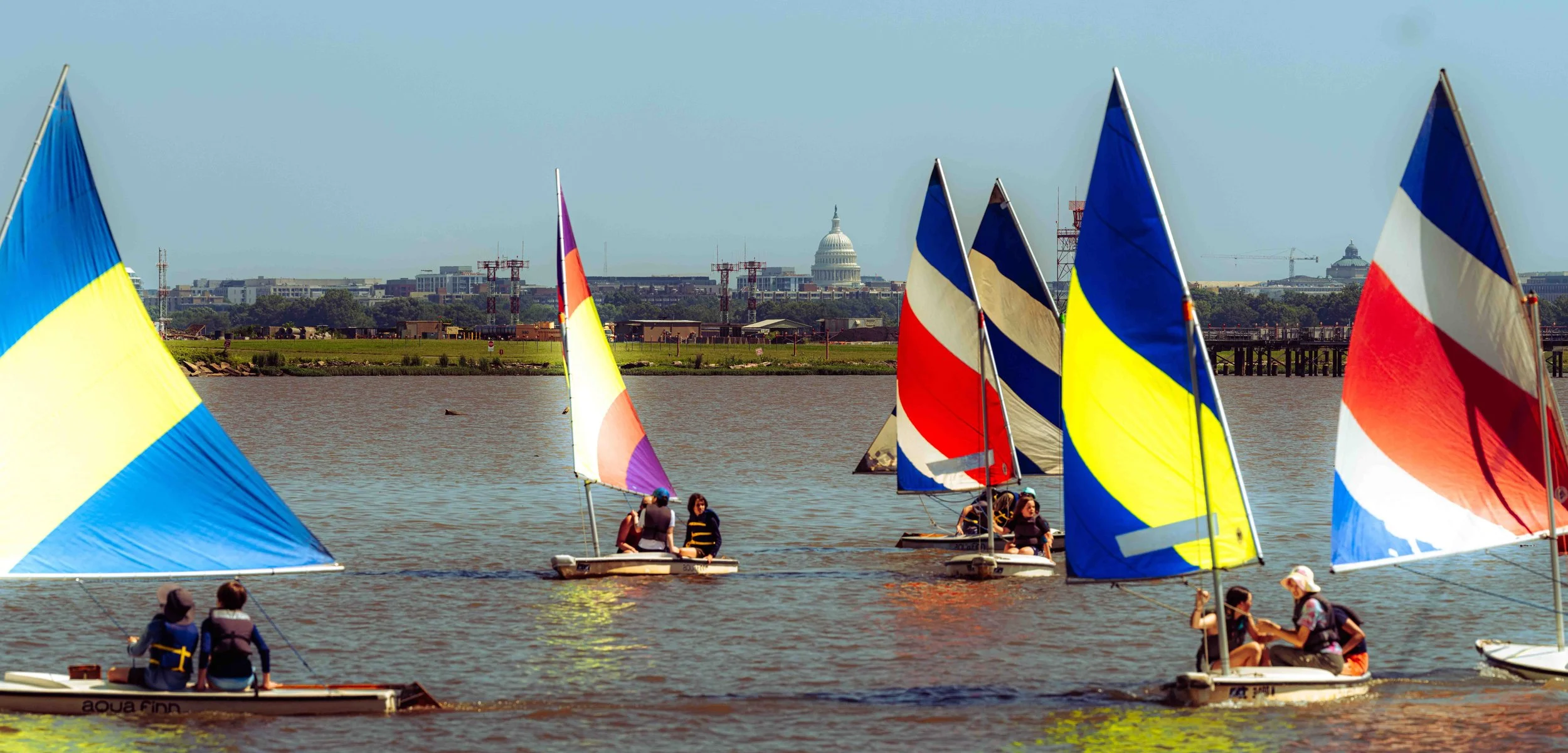 Student sailors navigate colorful dinghies on the Potomac River with the U.S. Capitol visible in the distance.